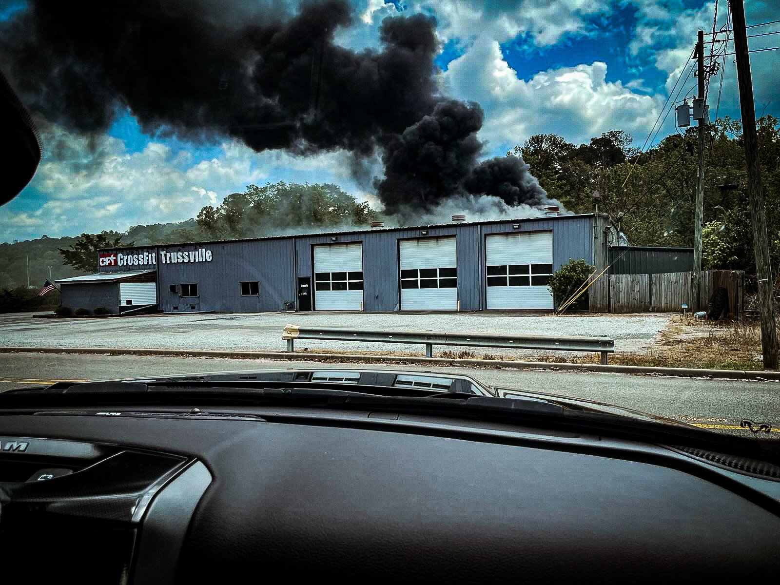 Fire and black smoke coming from a CrossFit gym building labeled 'CrossFit Trussville' with three garage doors, viewed from inside a vehicle.
