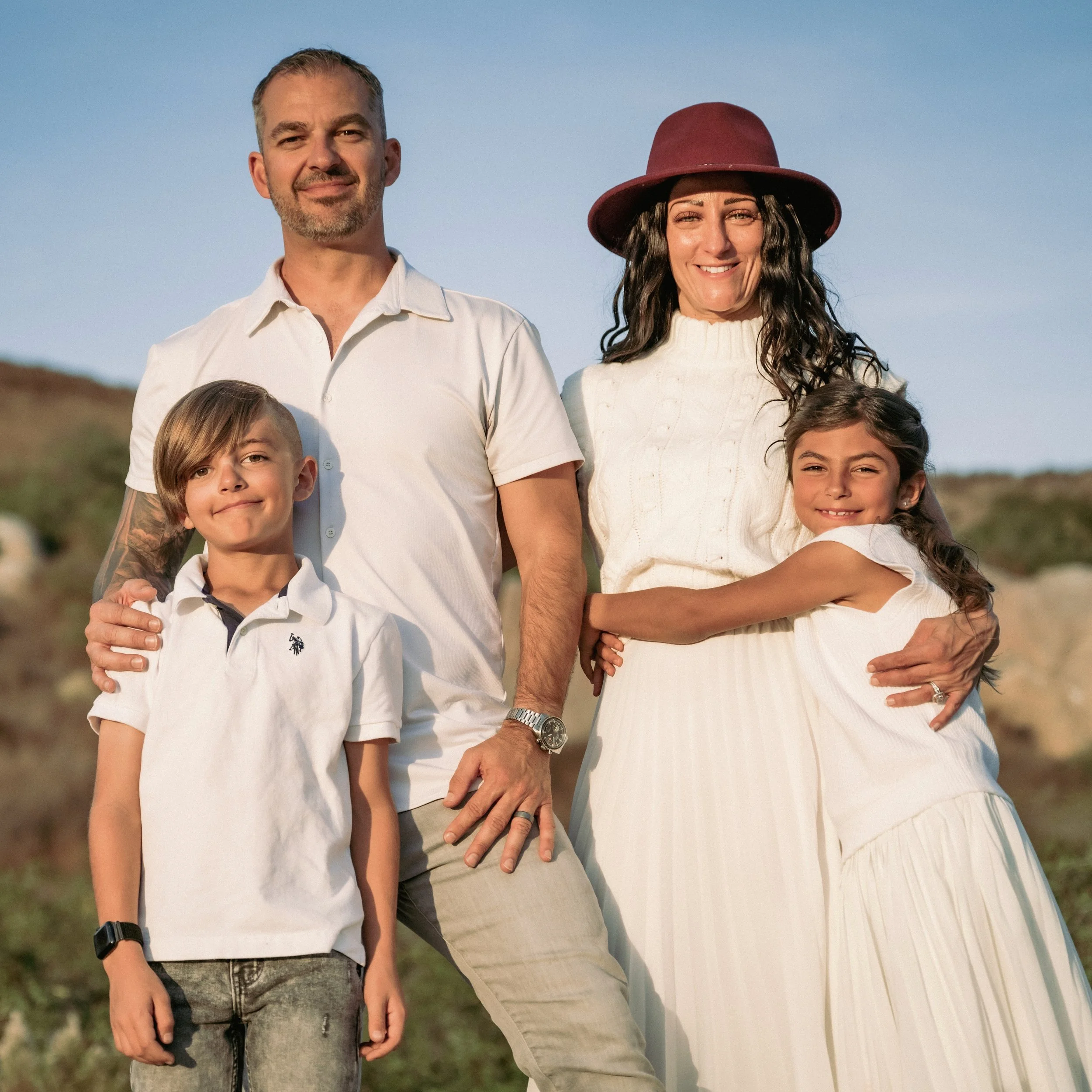 A group of people, including two children and two adults, standing outdoors against a clear blue sky. One adult woman wears a large maroon hat and a white sweater, while the man beside her has short hair, a beard, and is wearing a white shirt. Two children are partially visible, one with short hair and the other with long hair, all smiling and enjoying the moment.
