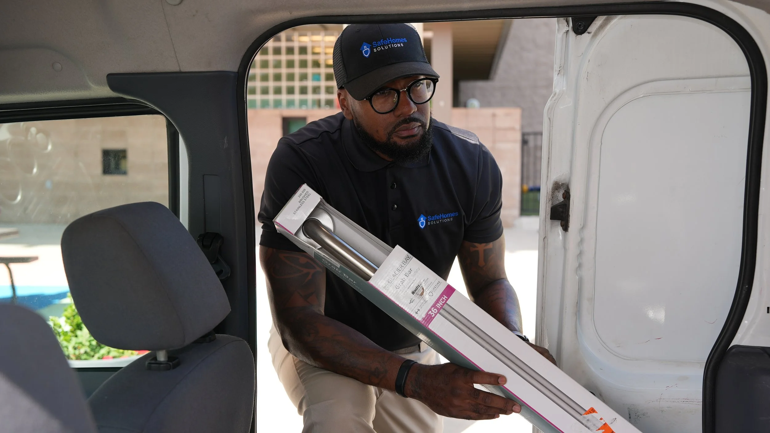 A man wearing glasses, a cap, and a dark polo shirt with a 'Safe Homes Solutions' logo, loading a boxed item into a vehicle.