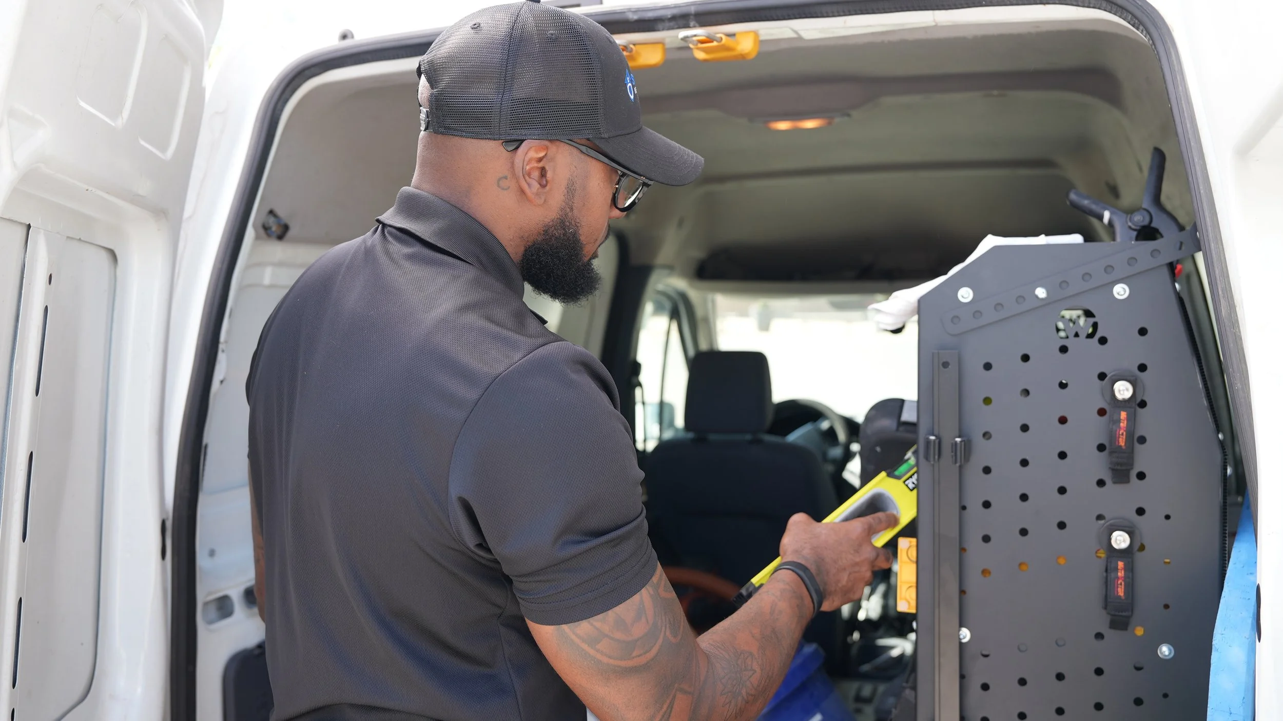 A technician wearing a black cap, glasses, and a gray polo shirt is working inside a vehicle, using a yellow testing or measurement tool on an open electronic panel.