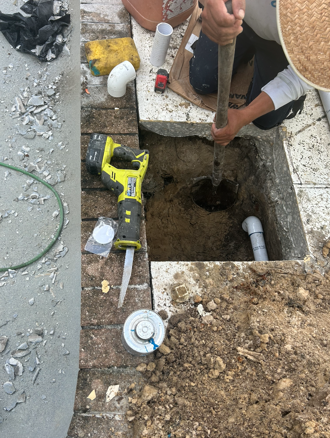A person installing or repairing underground plumbing pipe using a shovel in a construction site. Various tools and materials are scattered around, including a cordless saw, pipes, and a container of paint.