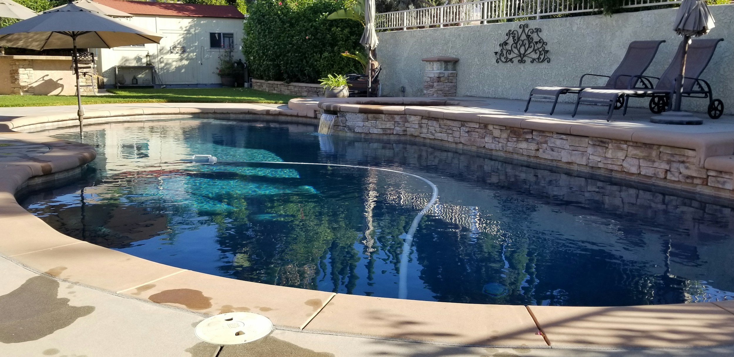 A backyard swimming pool with a waterfall feature, surrounded by a stone deck and lounge chairs. Pool cleaning equipment is visible in the water, and an umbrella provides shade nearby. There is a grassy area and a white fence in the background.