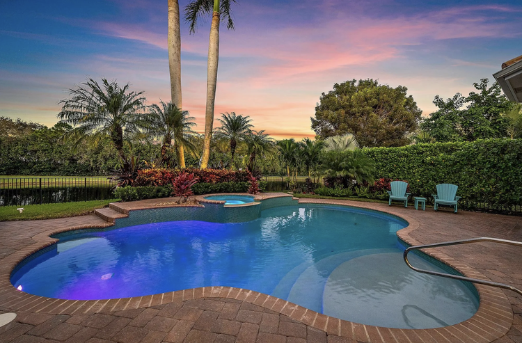 A backyard swimming pool with colorful lighting at sunset, surrounded by lush tropical plants, palm trees, and two blue chairs.