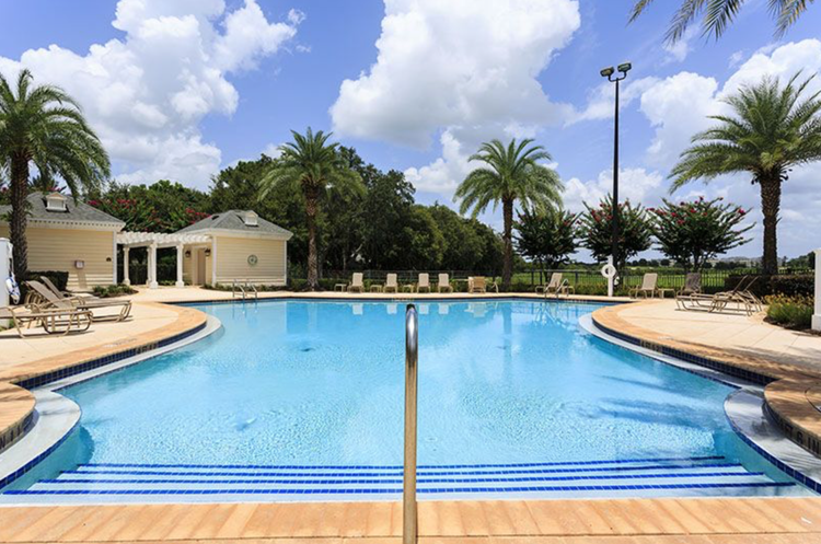 A resort swimming pool area with palm trees, lounge chairs, and a small yellow pool house under a partly cloudy sky.