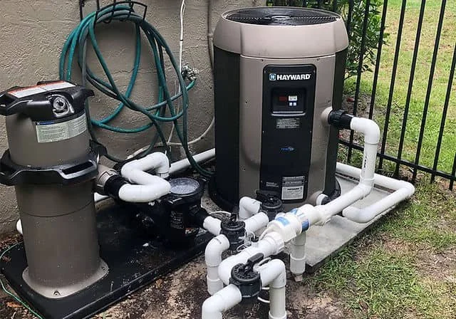 Pool filtration system with a Hayward sand filter, pool pump, and plumbing pipes outside on a concrete pad next to a fence and grass.
