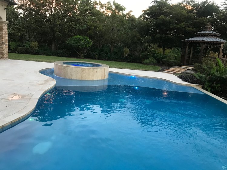 A backyard swimming pool with a raised hot tub, surrounded by a concrete deck, lush greenery, and a wooden gazebo in the background at sunset.