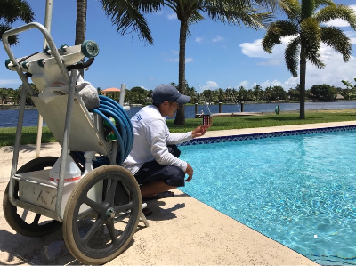 A man crouching by a swimming pool, taking a photo with his phone, with a pool cleaning cart nearby and palm trees in the background.