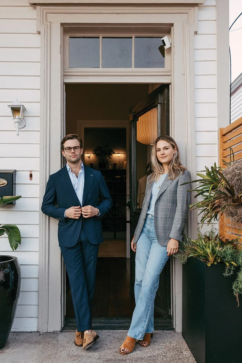 A man and woman standing at the entrance of a house, with the man wearing a blue suit and glasses, and the woman wearing a gray plaid blazer and light blue jeans, surrounded by potted plants.