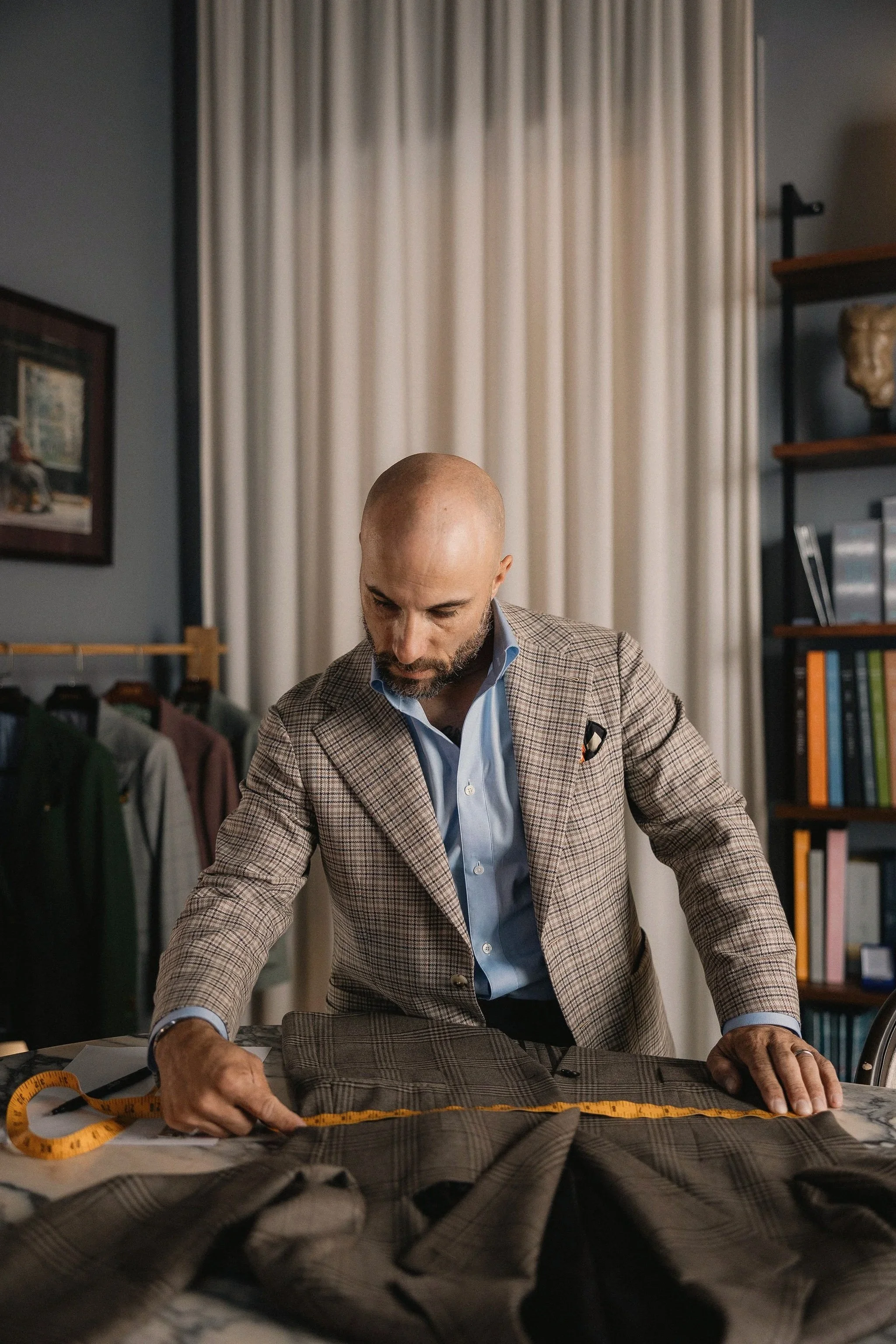 A man with a bald head, beard, and a checkered blazer working on a sewing project in a room with bookshelves. He is measuring fabric on a table with a measuring tape and fabric, surrounded by sewing supplies.