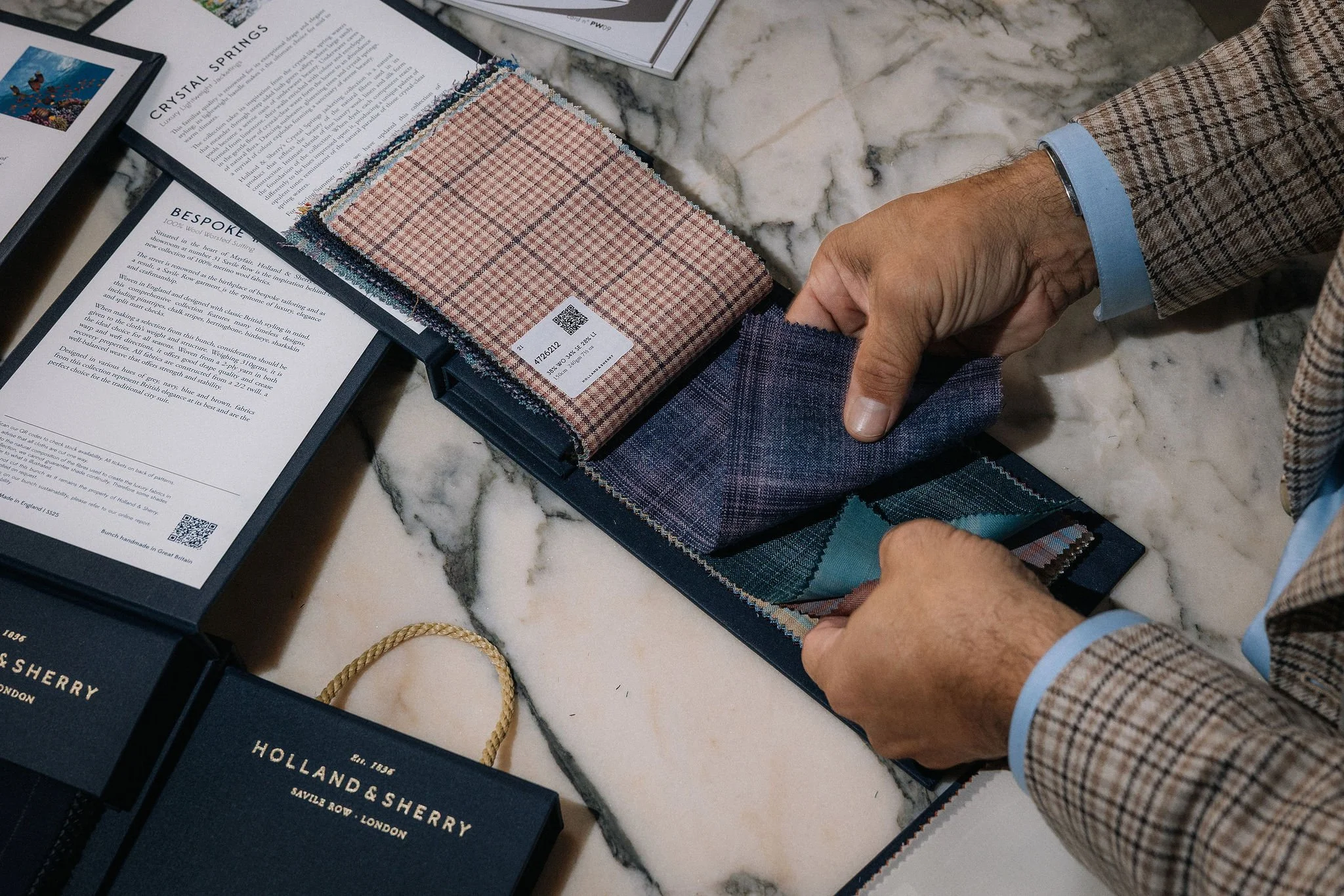 Person in a plaid blazer holding fabric swatches on a marble table, with fabric samples, brochures, and a QR code visible.