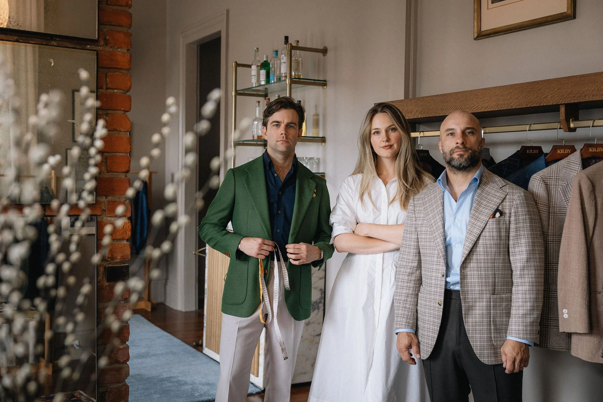Three stylish adults standing in a clothing boutique, with one man holding a measuring tape, surrounded by clothing on racks.