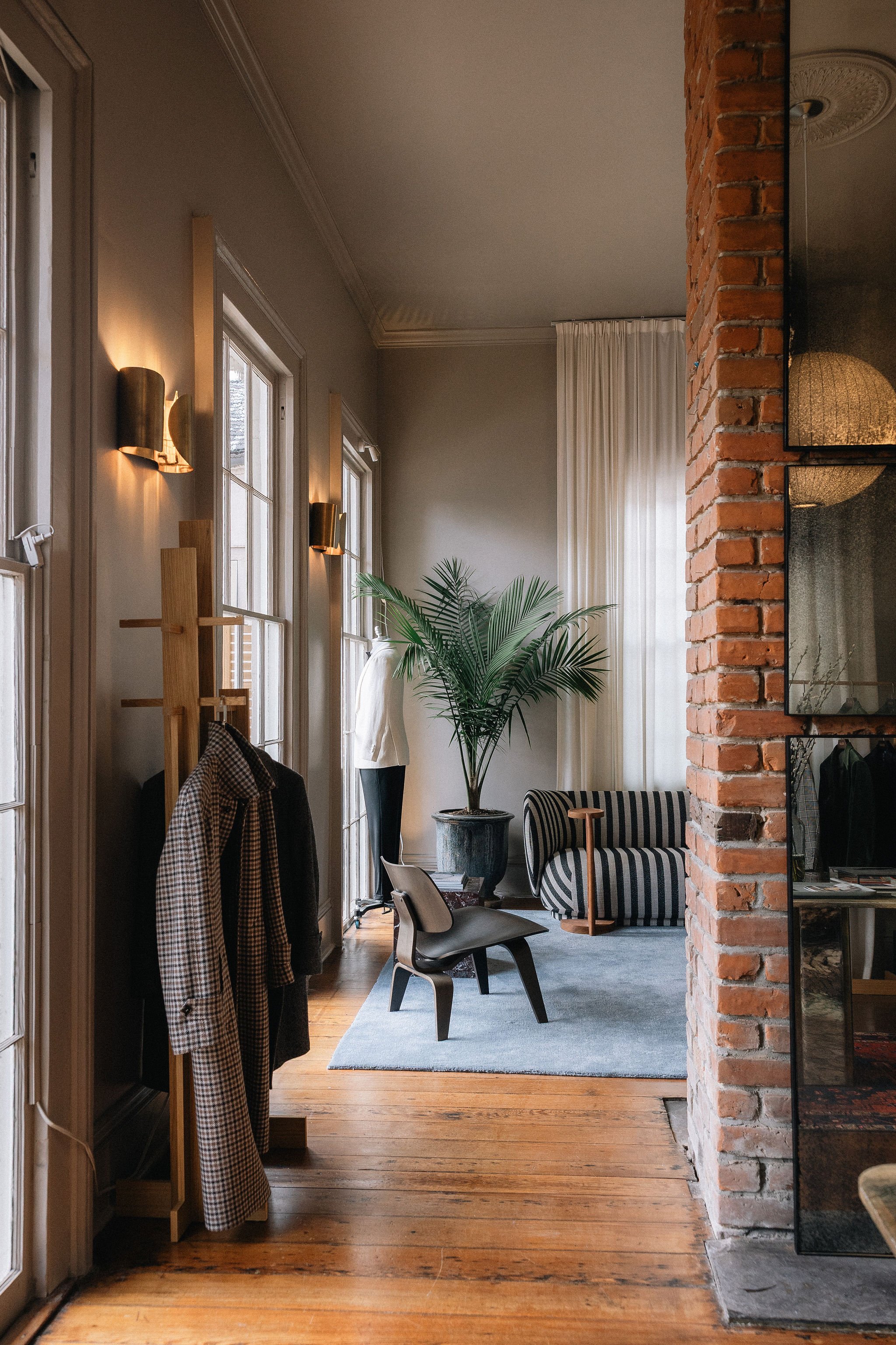 Interior of a living space with wooden floors, a brick wall, and large windows with white curtains. There is a green potted plant, a striped armchair, a modern chair, and clothing hanging on a wooden rack. Wall-mounted lights illuminate the wall, and a mirror reflects part of the room.