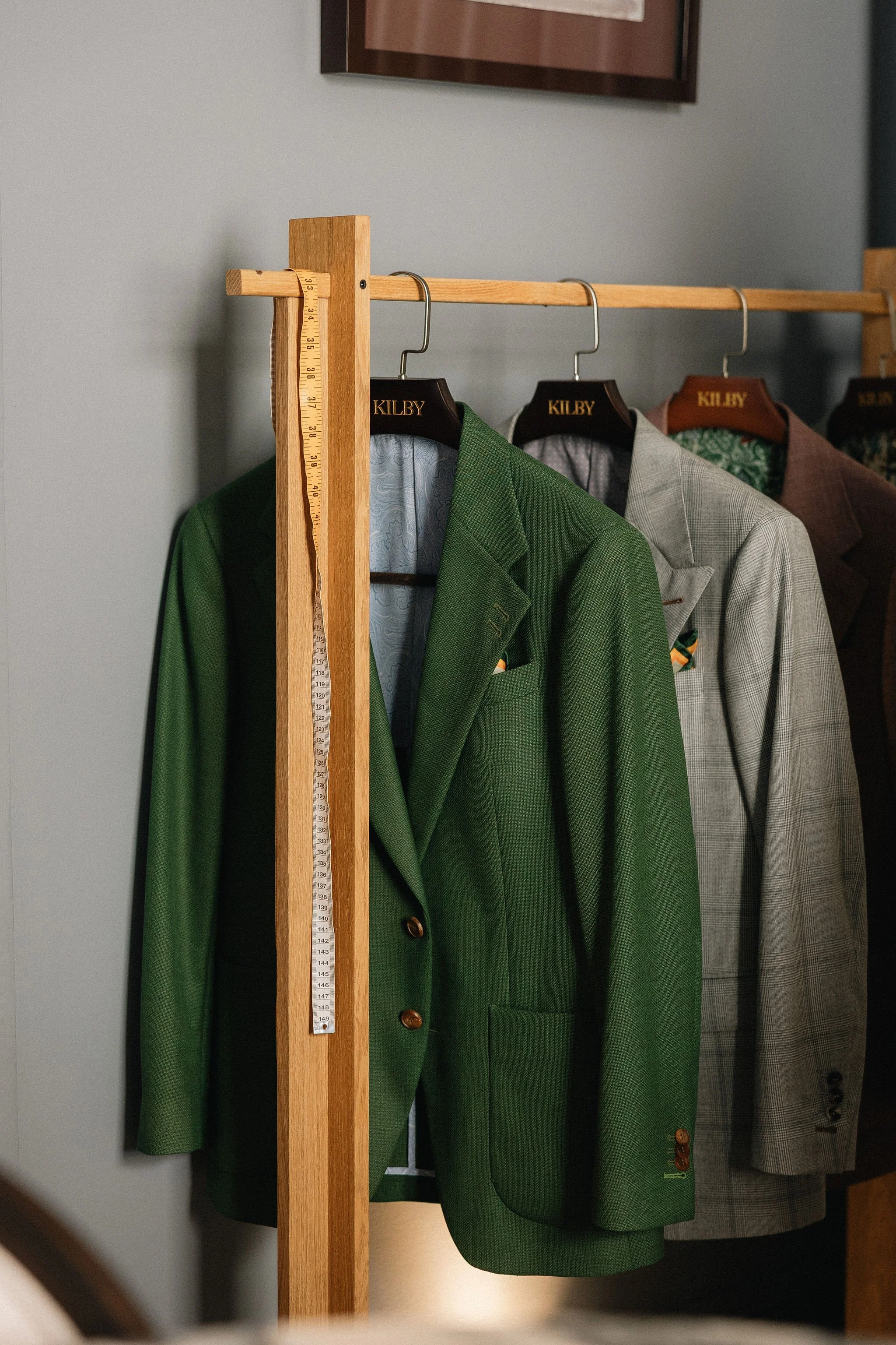 A wooden clothing rack with suits hanging on it, including a green suit, a gray suit, and a brown suit. The suits are on hangers labeled 'KILBY' and the rack is aligned against a wall with a picture frame above.