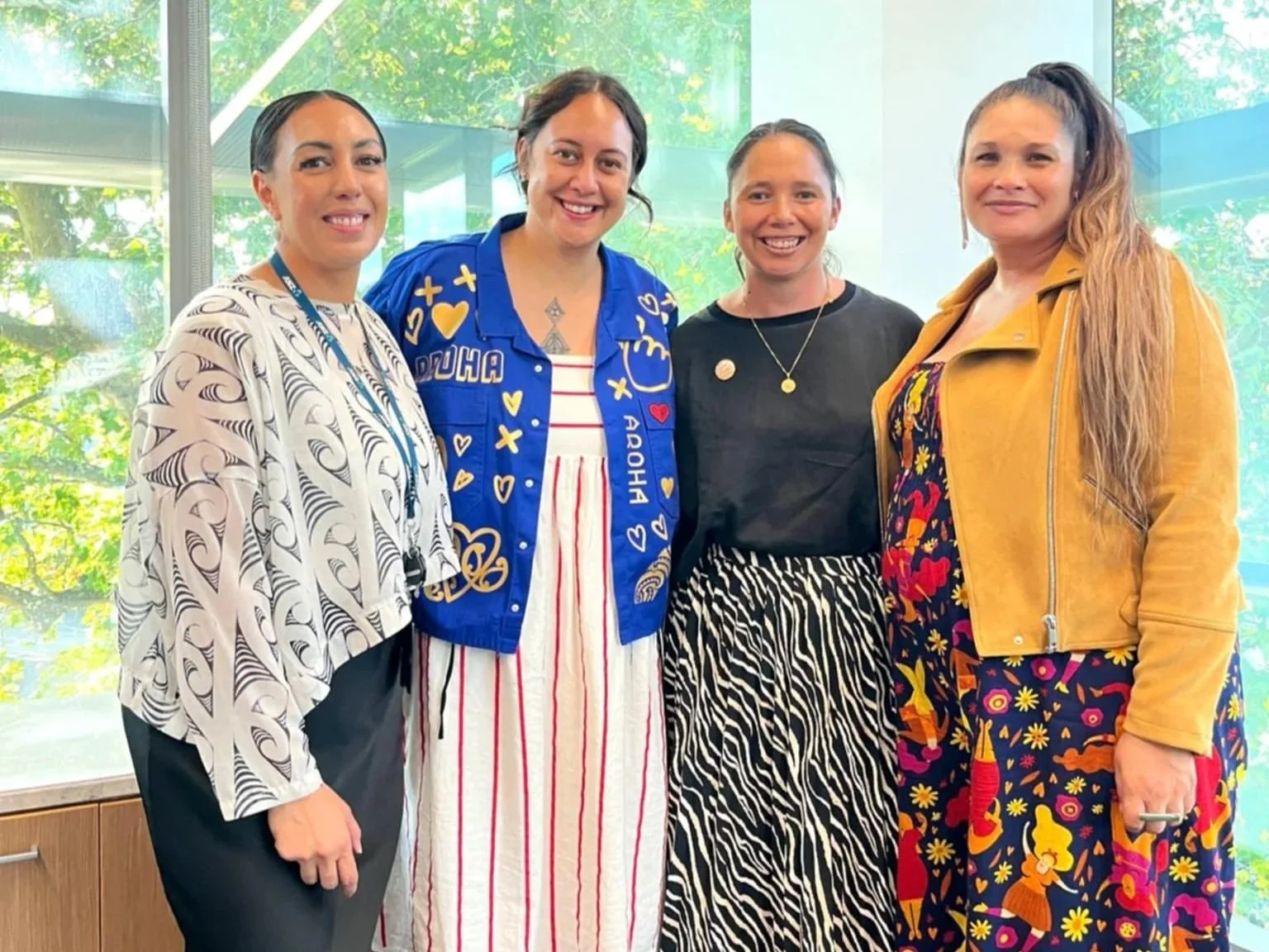 Four women standing together indoors with a large window behind them showing trees and sunlight. They are smiling and dressed in colorful, patterned attire.
