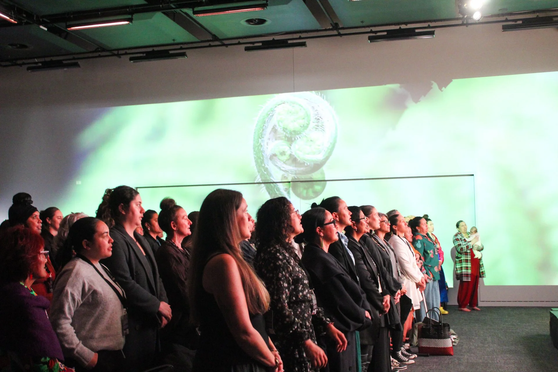Group of diverse women standing and watching a presentation or performance in a dark room with a large screen displaying nature imagery in the background at Te Mana o Ngā Wāhine.