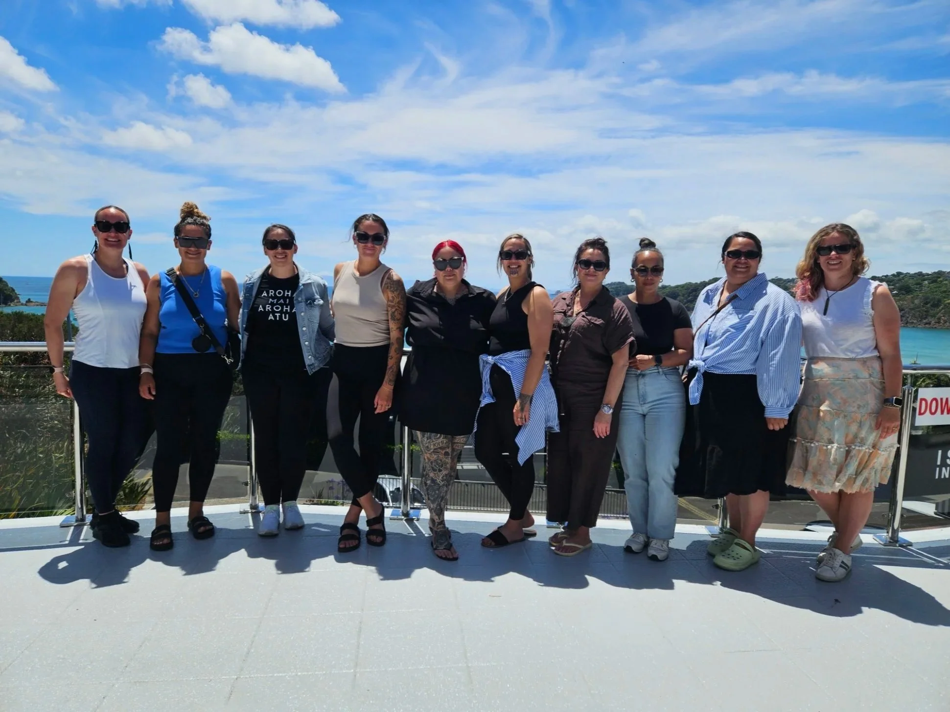 A group of ten women standing together outdoors on a balcony with ocean and green hills in the background, wearing casual summer clothing and sunglasses.