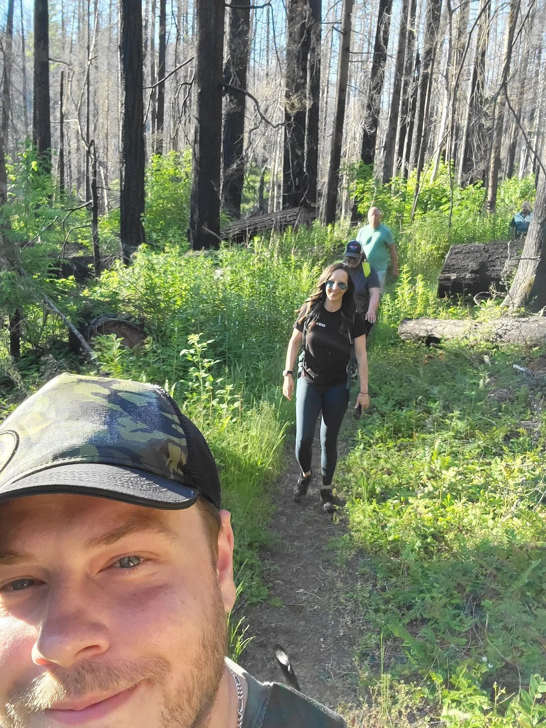 Group of people hiking on a forest trail, surrounded by tall trees and green foliage, with a woman smiling at the camera.