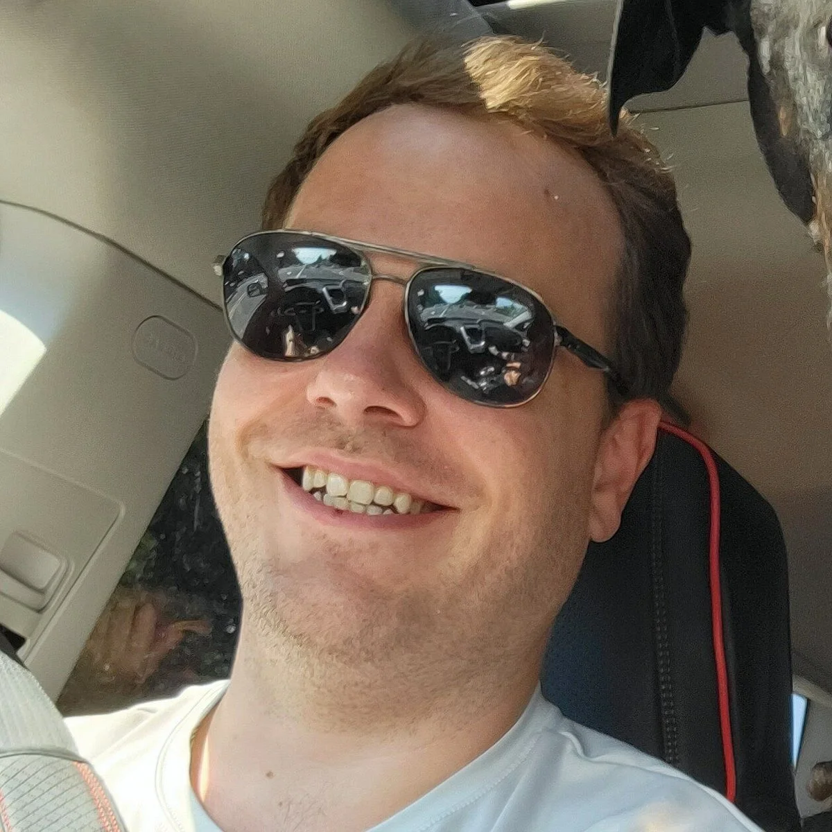 A man with short brown hair wearing sunglasses and smiling, sitting in a vehicle.