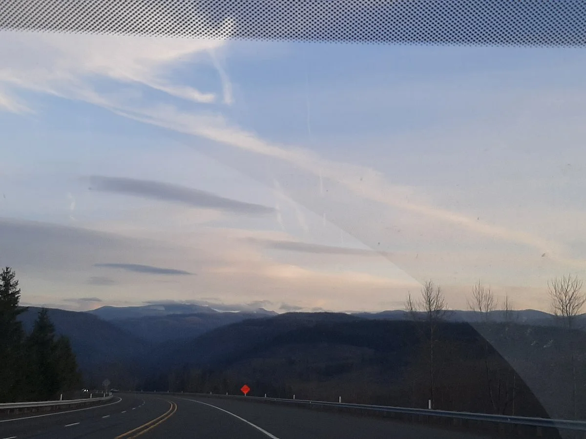 View of a winding mountain road seen through a car windshield with a partly cloudy sky and distant mountain range.