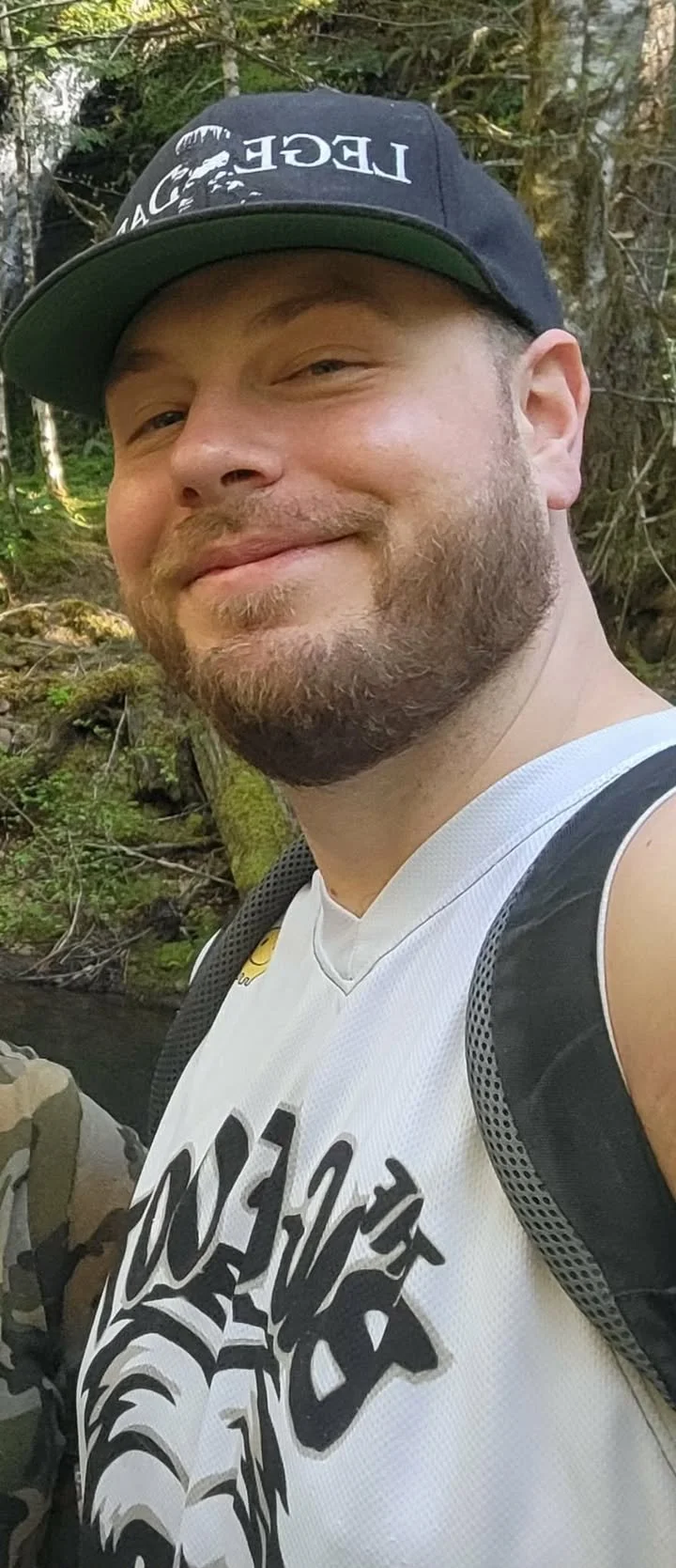 A smiling man with a beard wearing a black cap, a white graphic tank top, and a backpack outdoors in a forested area.
