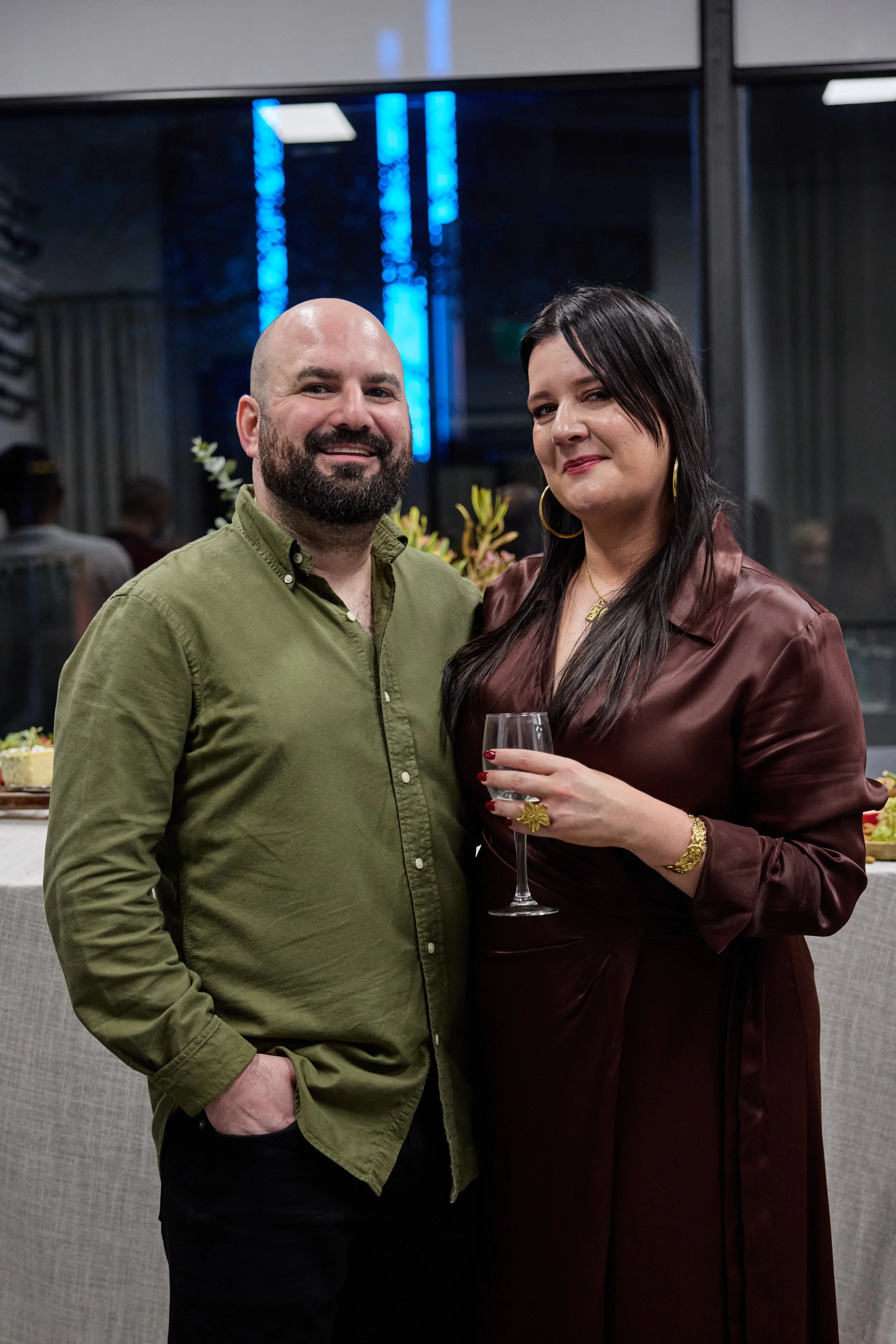 A man and woman posing together at an indoor event, with the woman holding a glass of champagne. The man is wearing a green button-up shirt, and the woman is dressed in a maroon satin dress with gold jewelry, including hoop earrings, a necklace, a wa
