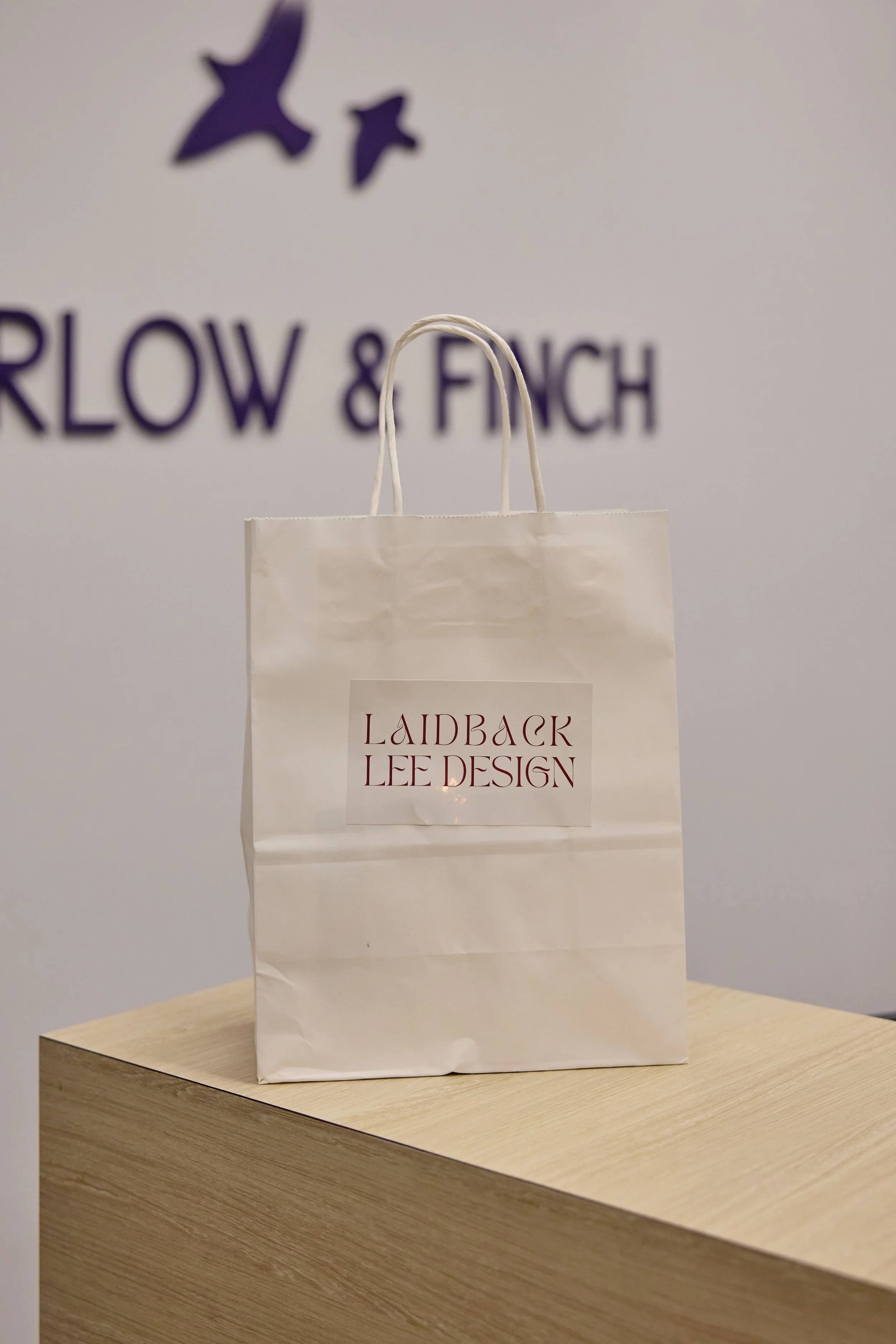 White shopping paper bag with a label reading "LAIDBACK LEE DESIGN" placed on a wooden surface. In the background, text on the wall reads "MARLOW & FINCH" with a logo of three stylized birds.