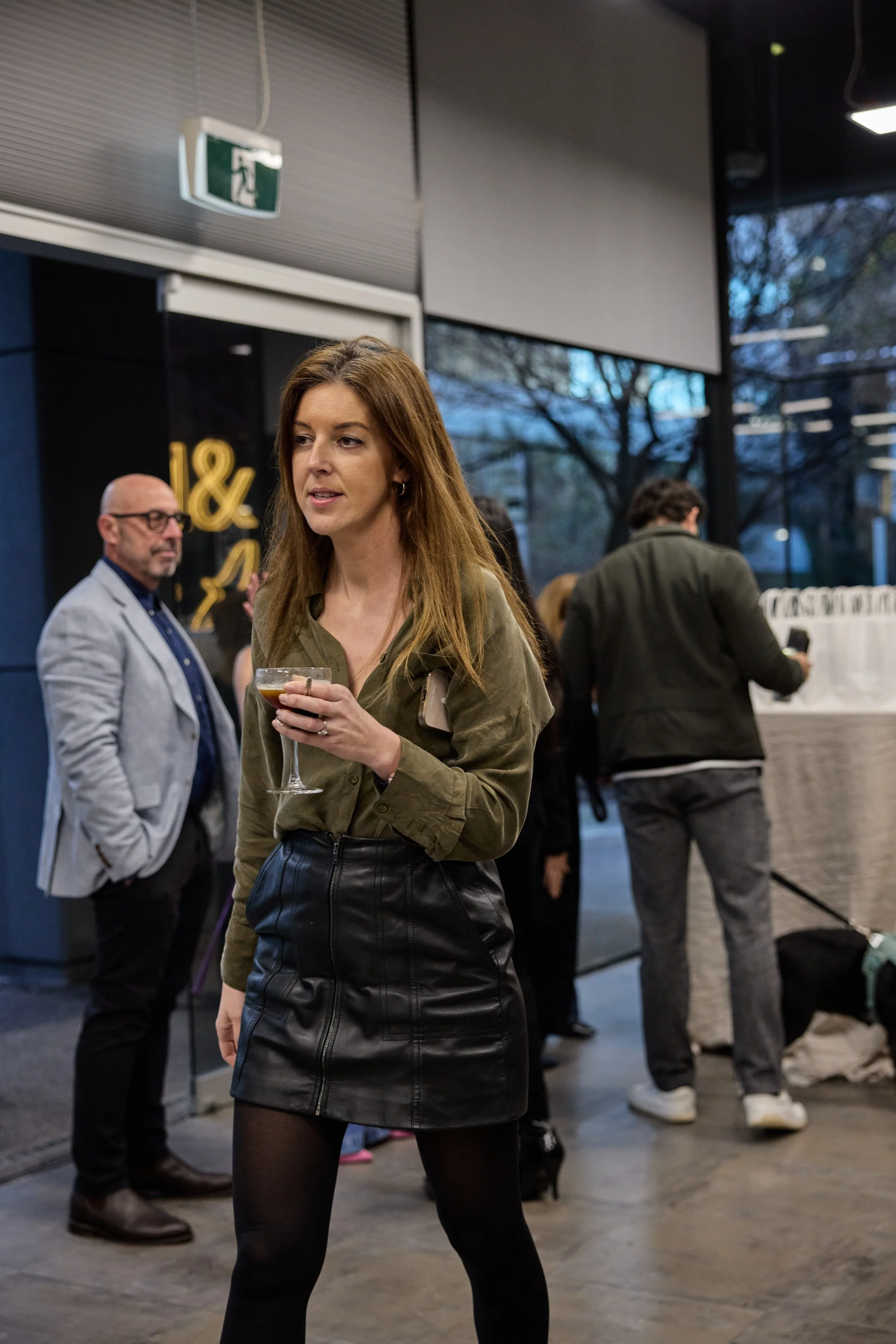 Woman in green shirt and black leather skirt holding a drink at an indoor social event, with people in the background near a large window.