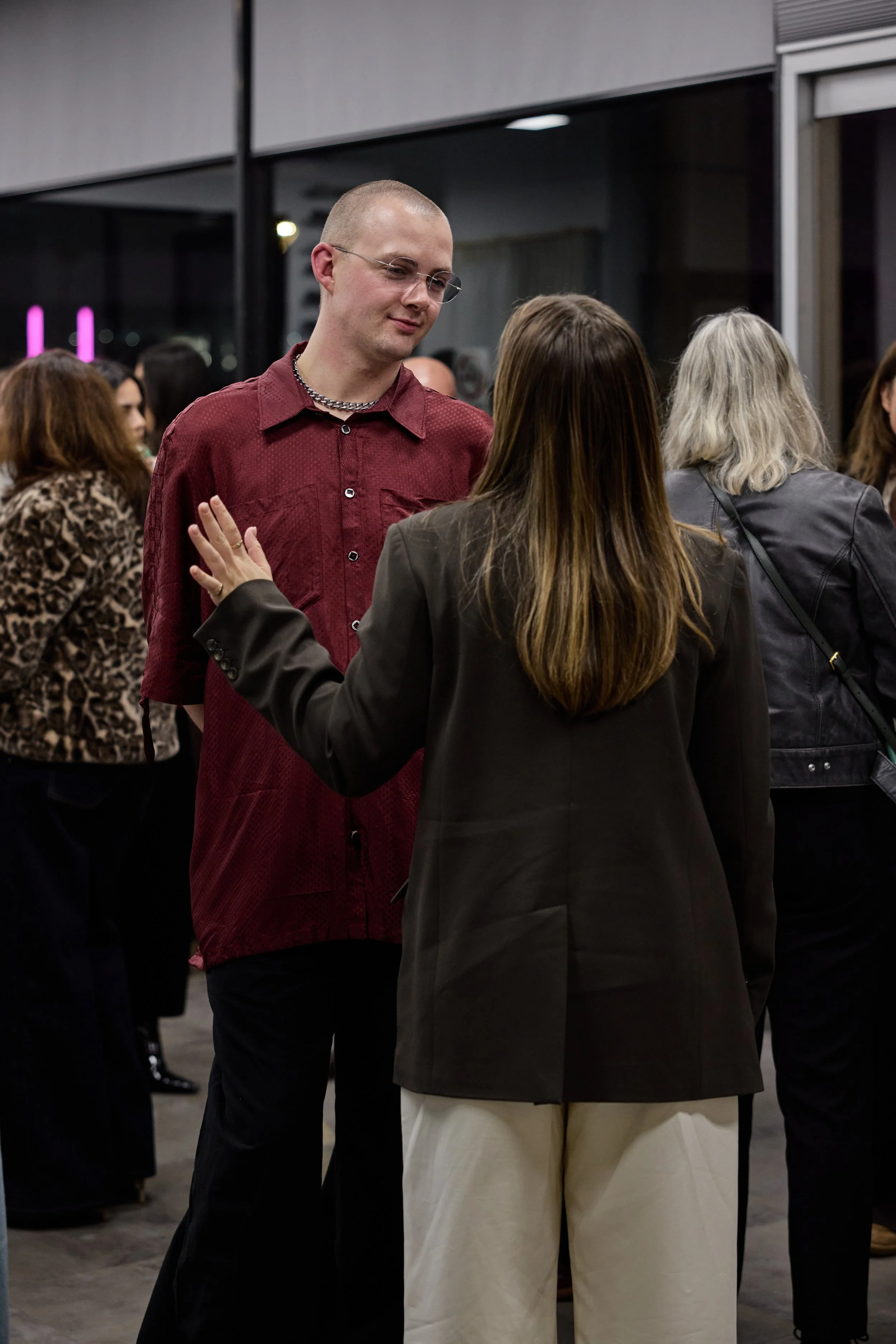 A man wearing glasses, a red shirt, and a necklace is speaking with a woman with long brown hair, wearing a black blazer and beige pants, in a crowded indoor setting.