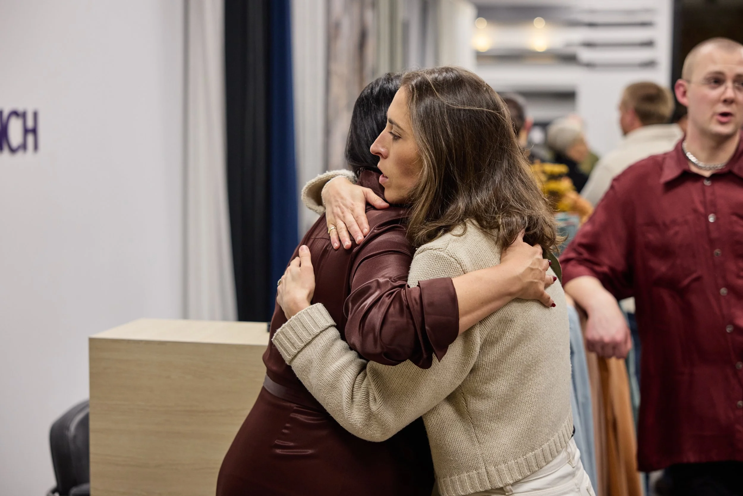 Two women hugging each other warmly in a social setting, with other people in the background.