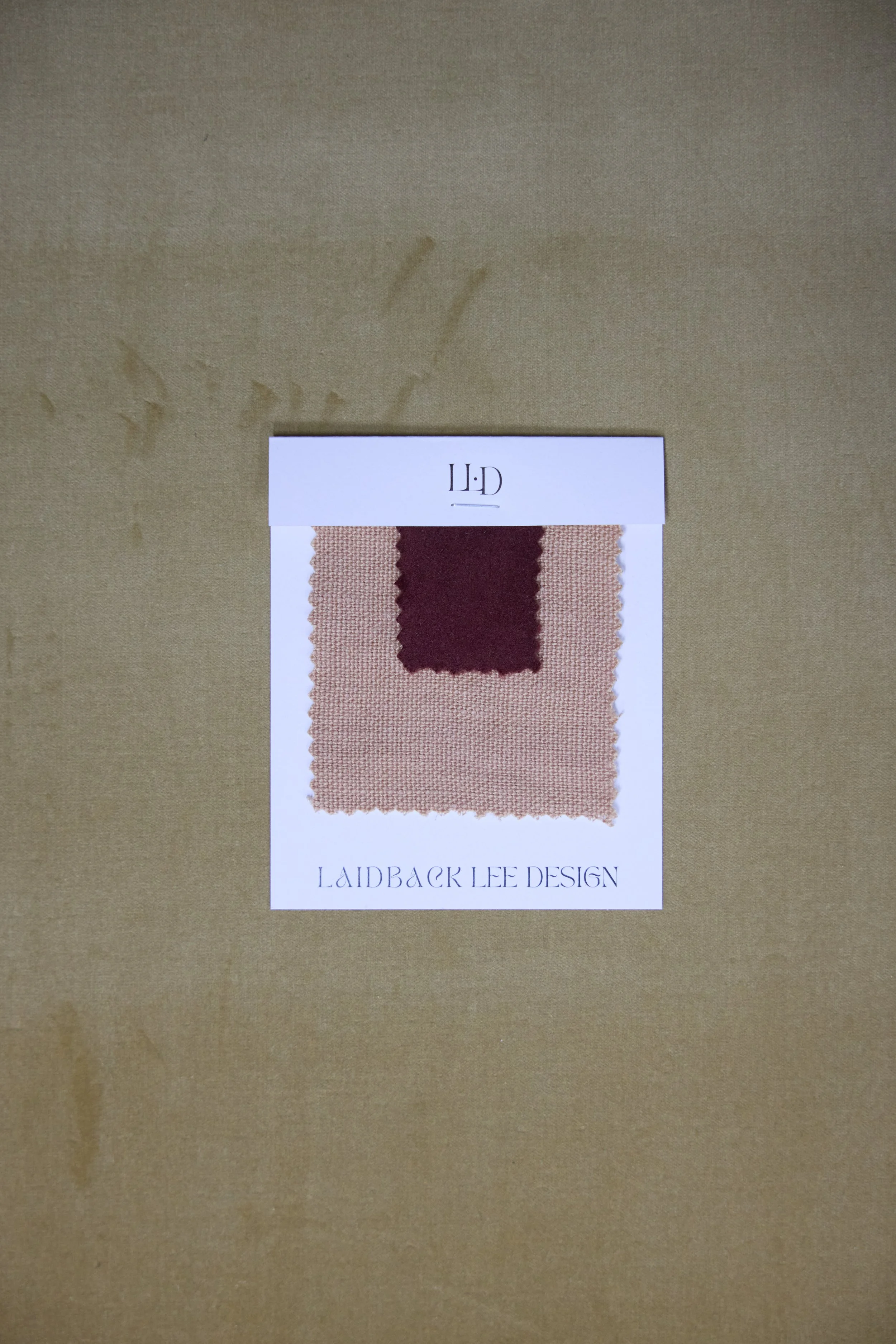Swatch card with two fabric samples, one burgundy and one beige, attached to a white backing card labeled 'Lailback Lee Design' on a brown surface.
