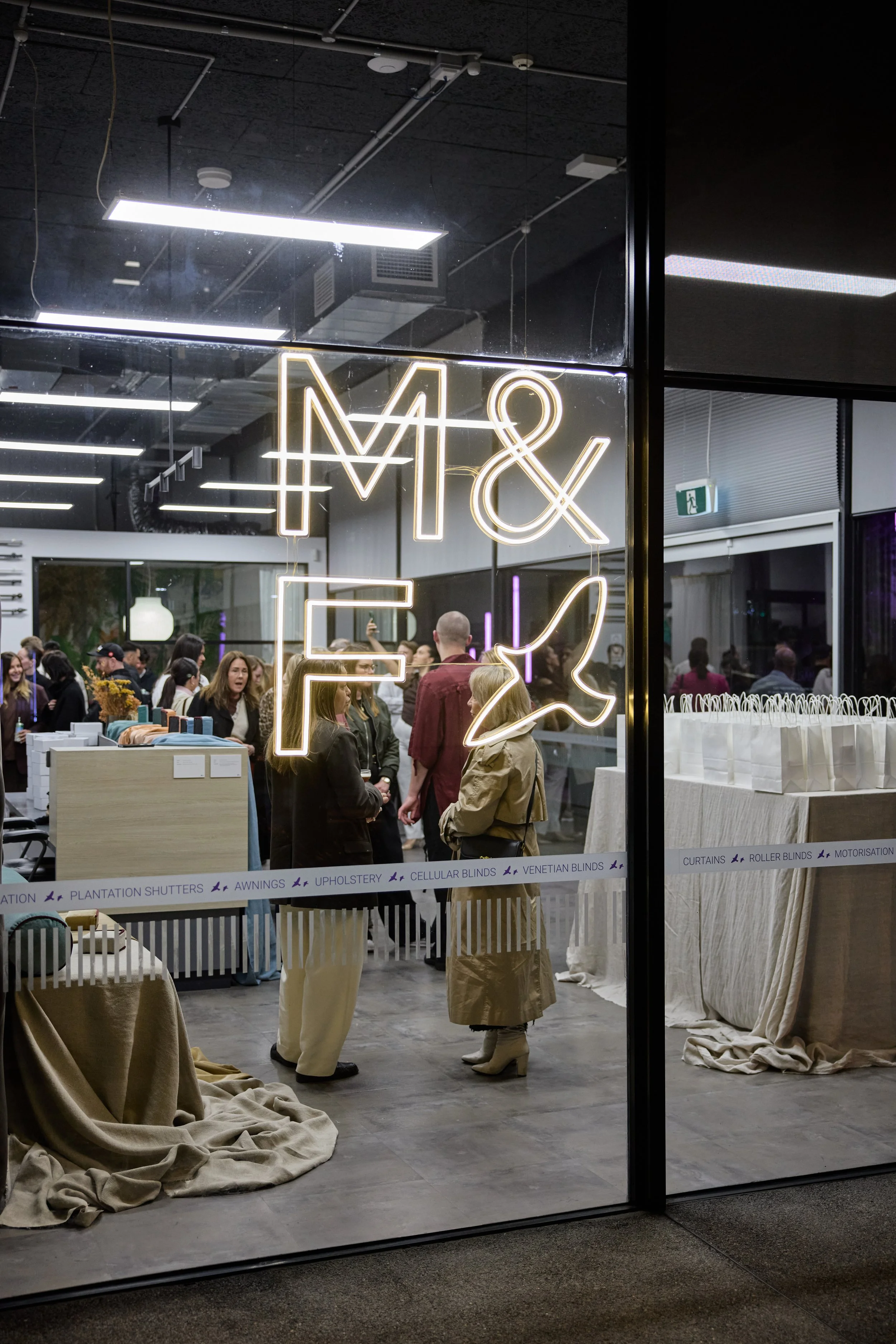 People gather inside a modern store or event space with a neon sign reading 'M & F' and a dove symbol. The store displays business related to curtains, blinds, upholstery, and related products, with shelves and tables covered in beige fabric and gift