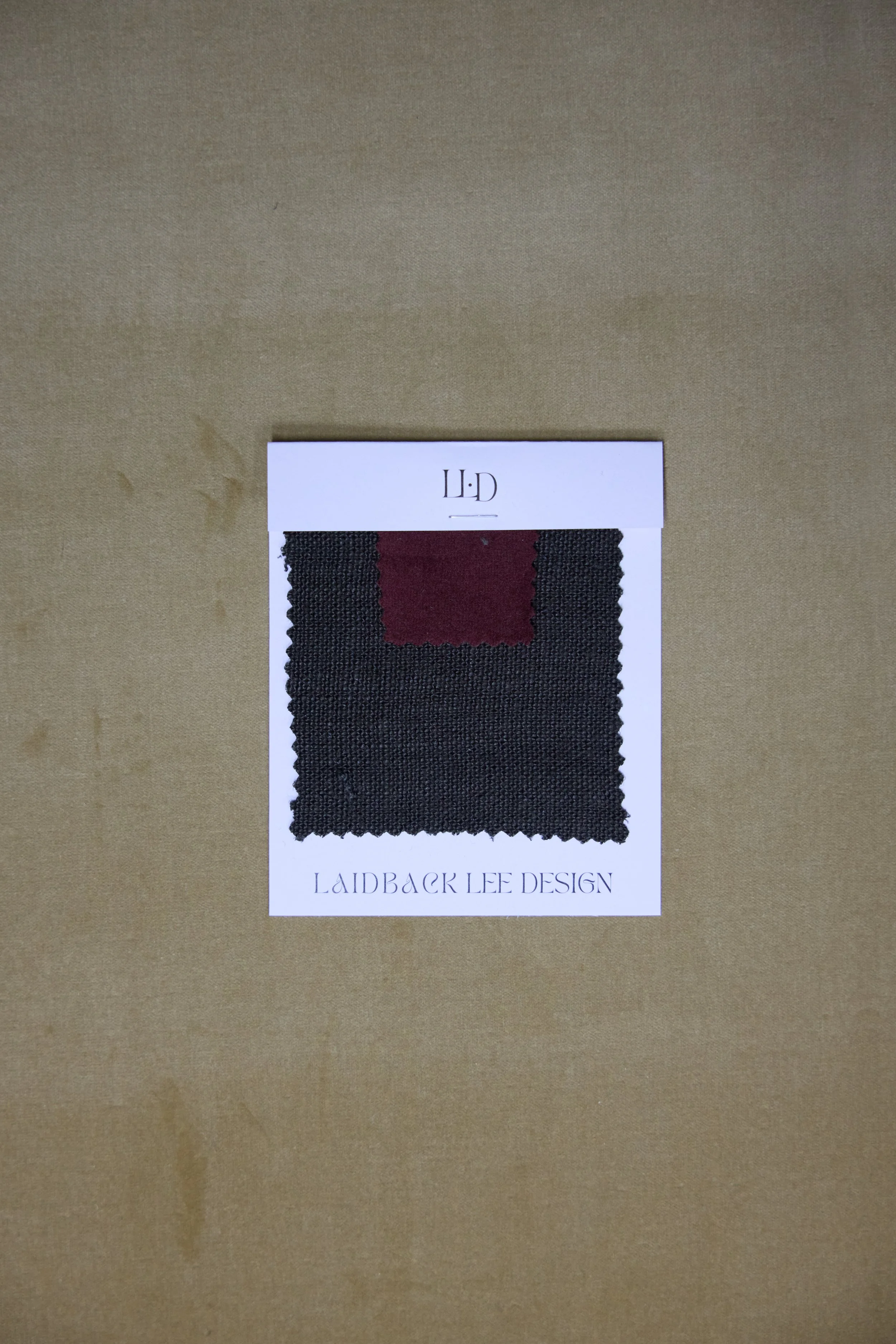 Fabric swatch in black with a small burgundy section, attached to a white card labeled 'Laidback Lee Design' on a tan surface.