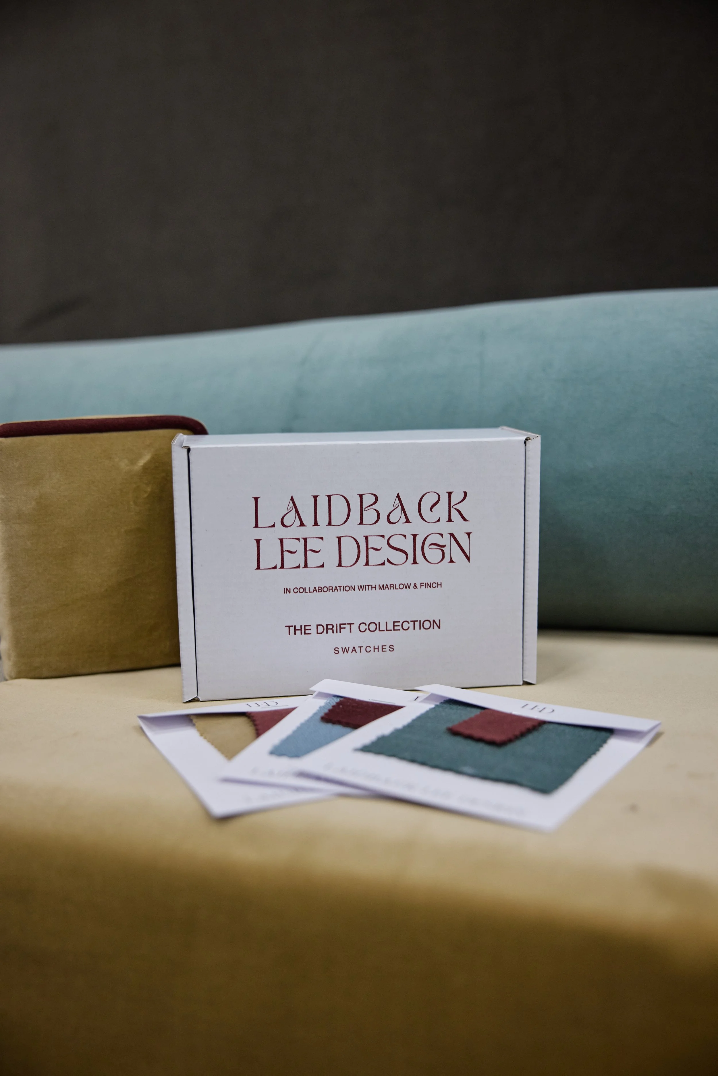 Swatches from the Laidback Lee Design Drift Collection displayed on a table with a box and a brown and blue bag in the background.