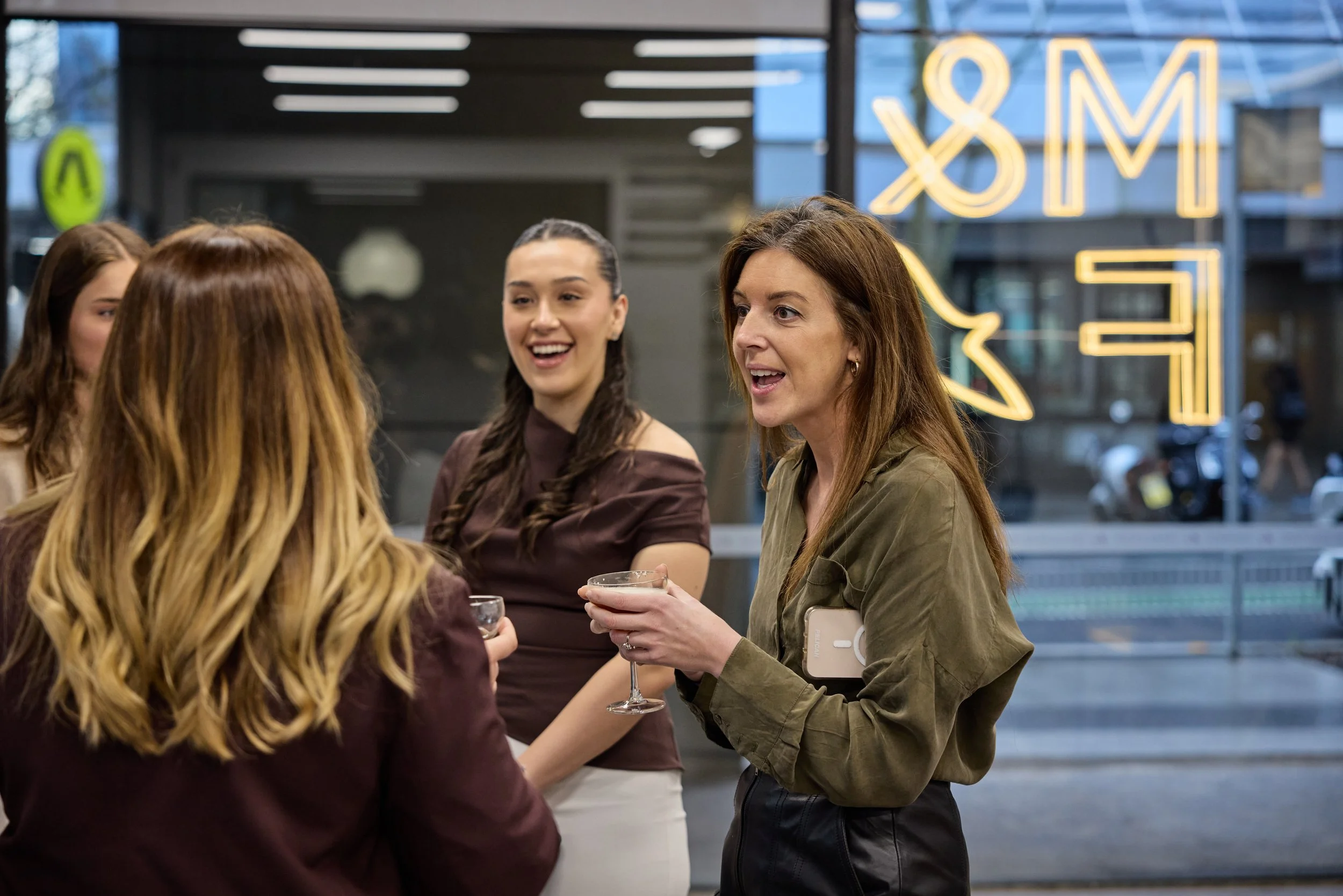 A group of women socializing with drinks in front of a neon sign in a modern indoor setting.
