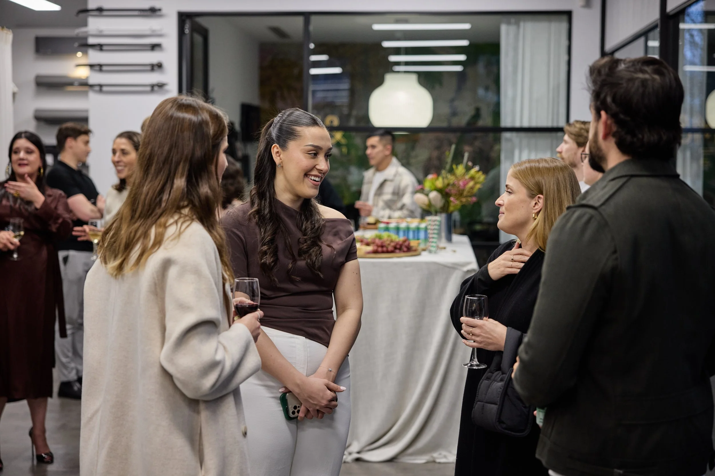 Group of people socializing at a party, holding wine glasses, indoors with a decorated table in the background.