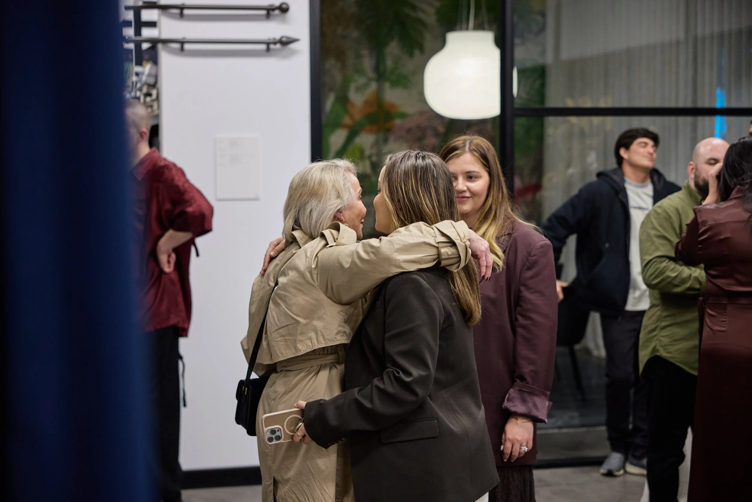 Three women standing close together, with one woman hugging another and the third woman looking on, in a social gathering. Others are seen in the background.
