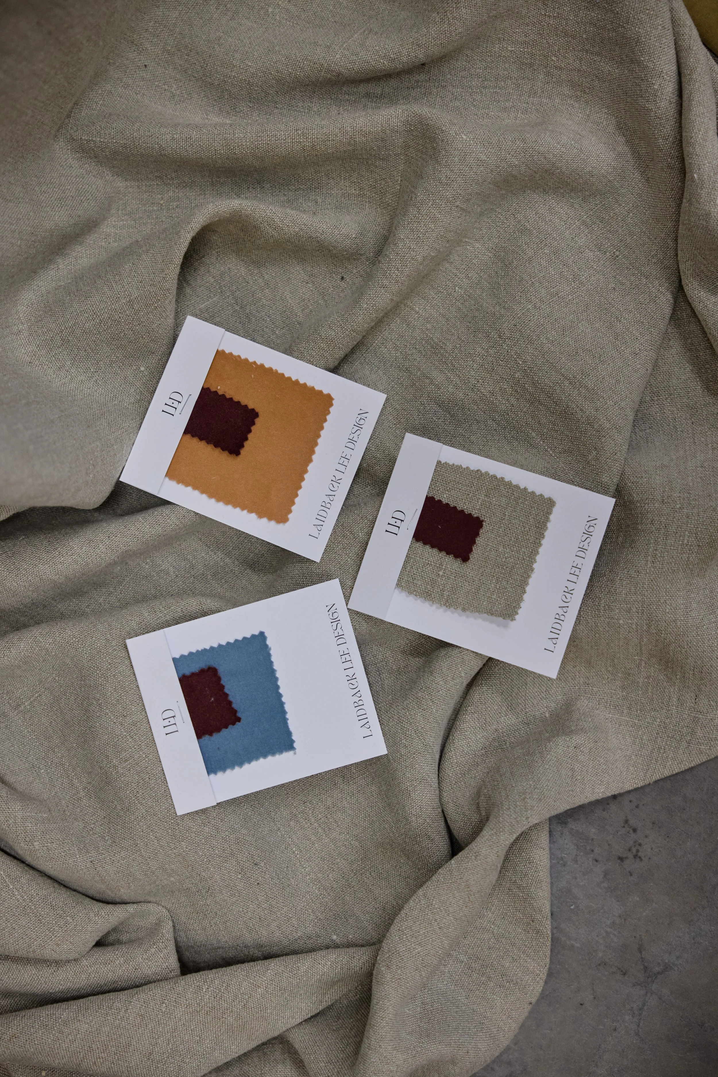 Three fabric swatch sample cards with different color combinations, placed on a beige fabric cloth.