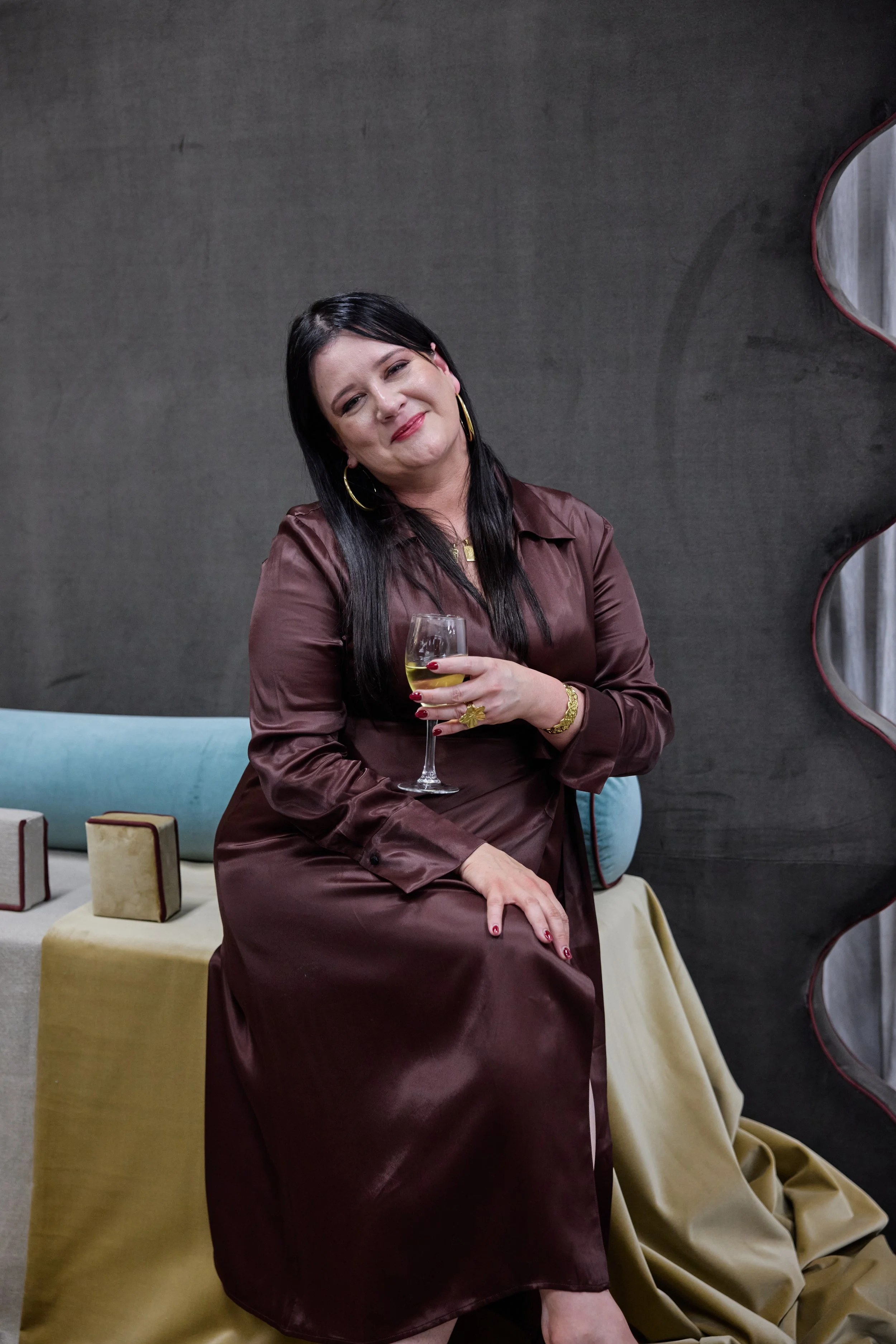 A woman with long black hair, wearing a shiny brown satin dress, holding a glass of white wine, sitting on a light-colored table with decorative boxes and against a gray wall.