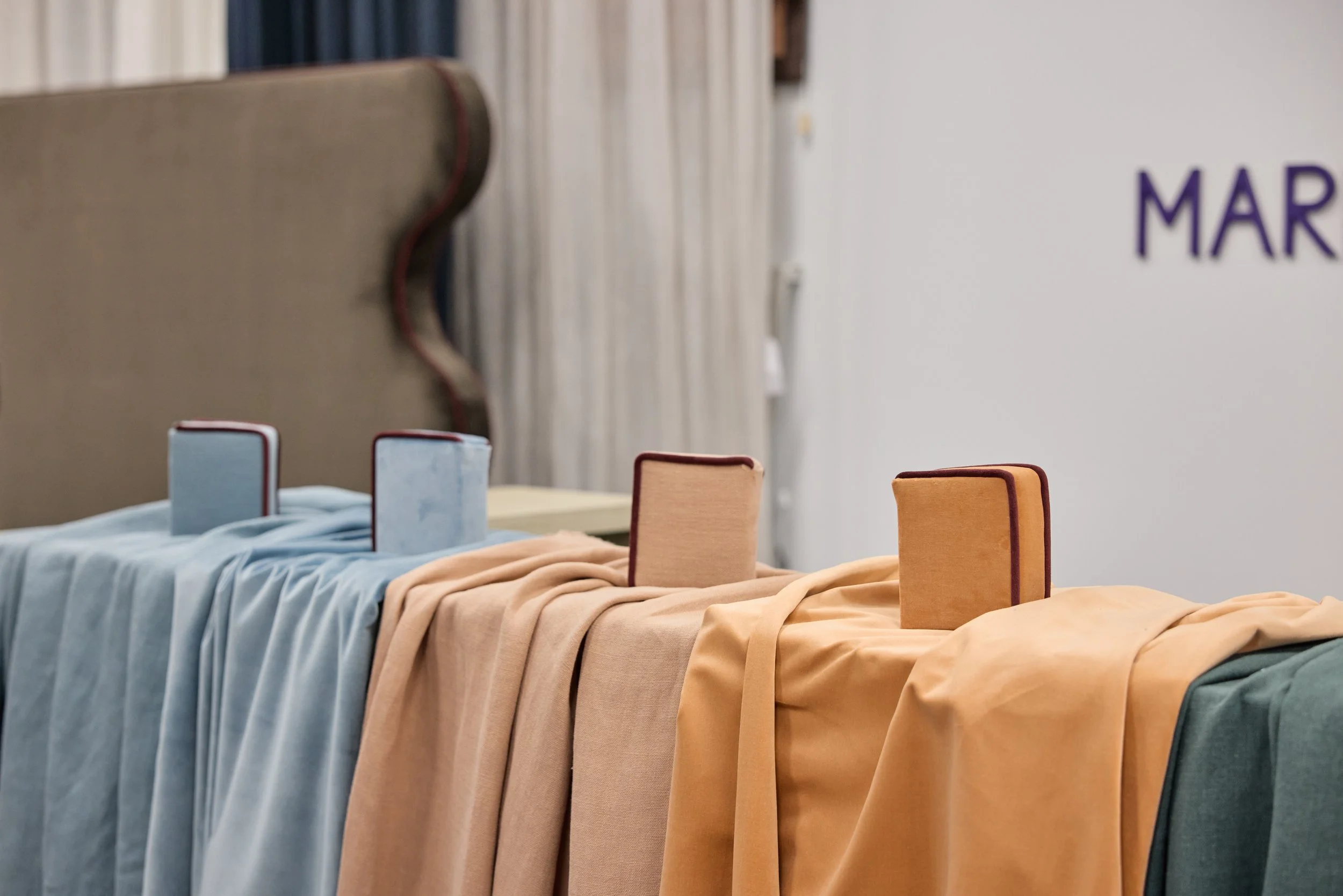 Pieces of fabric draped over a table with labeled books or boxes placed on top, set against a background with curtains and a white wall.