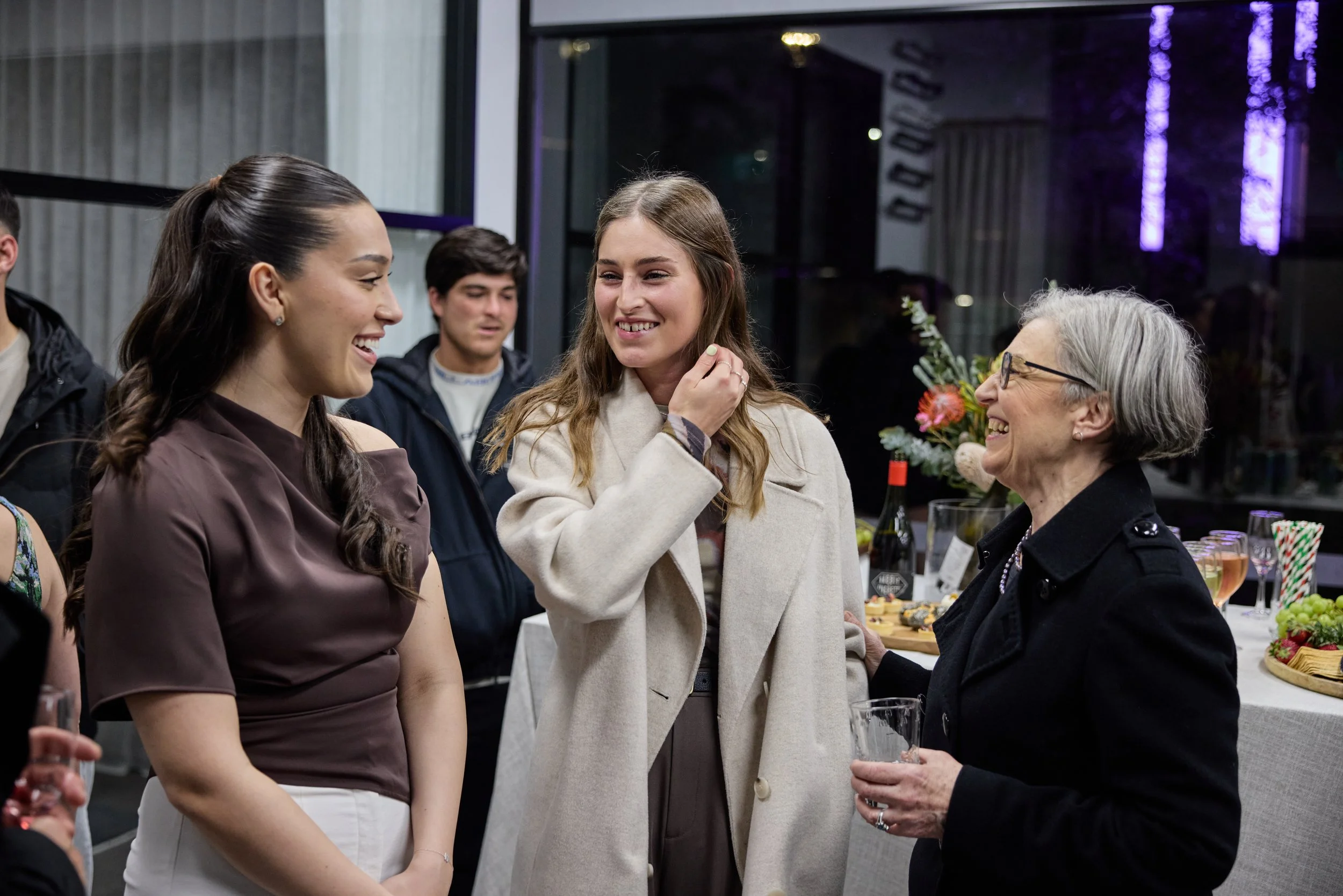 Group of women sharing a joyful moment at a social gathering, with a table of food and drinks in the background.