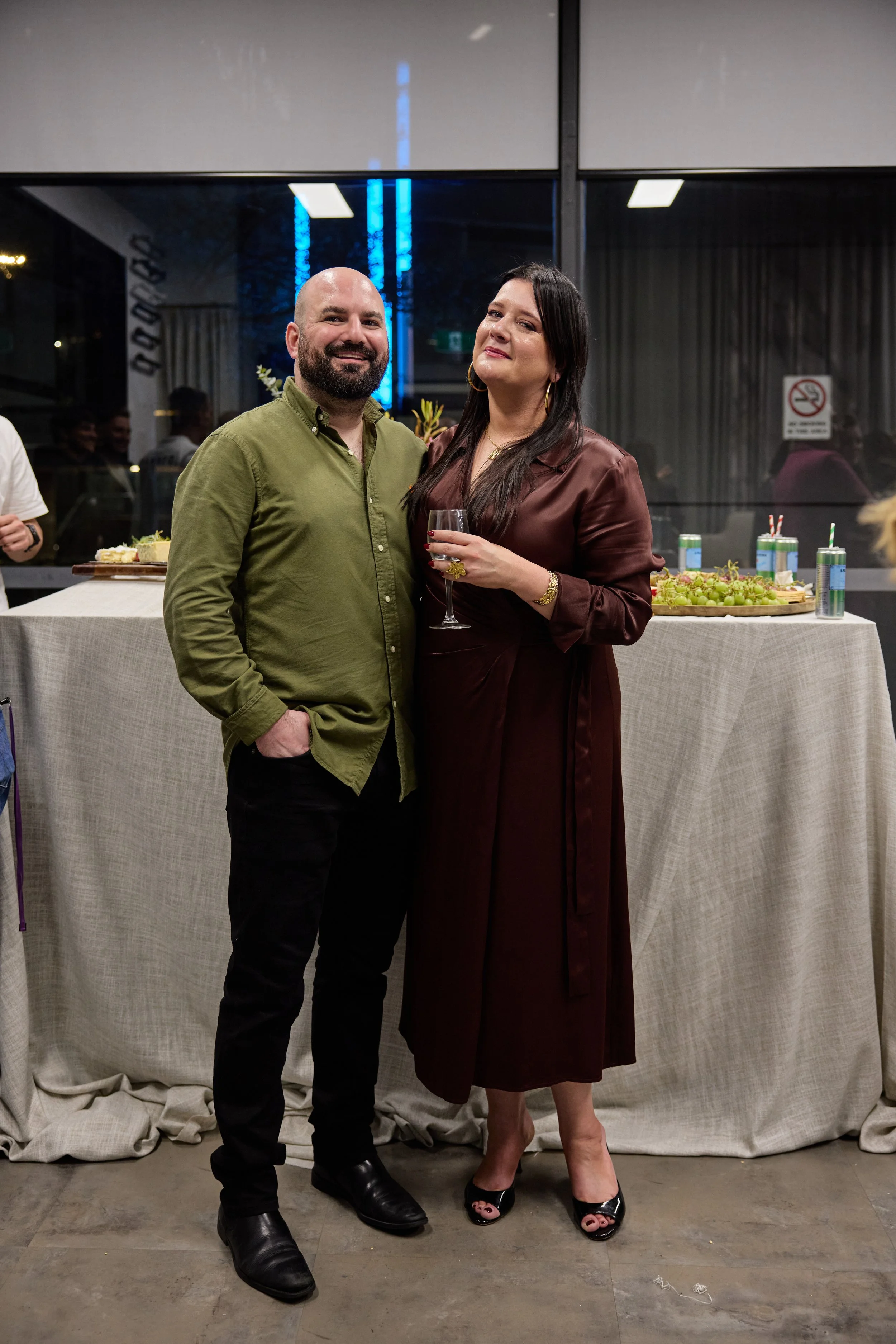 A man and woman standing together inside a building on a social occasion, with a table of snacks and drinks behind them.