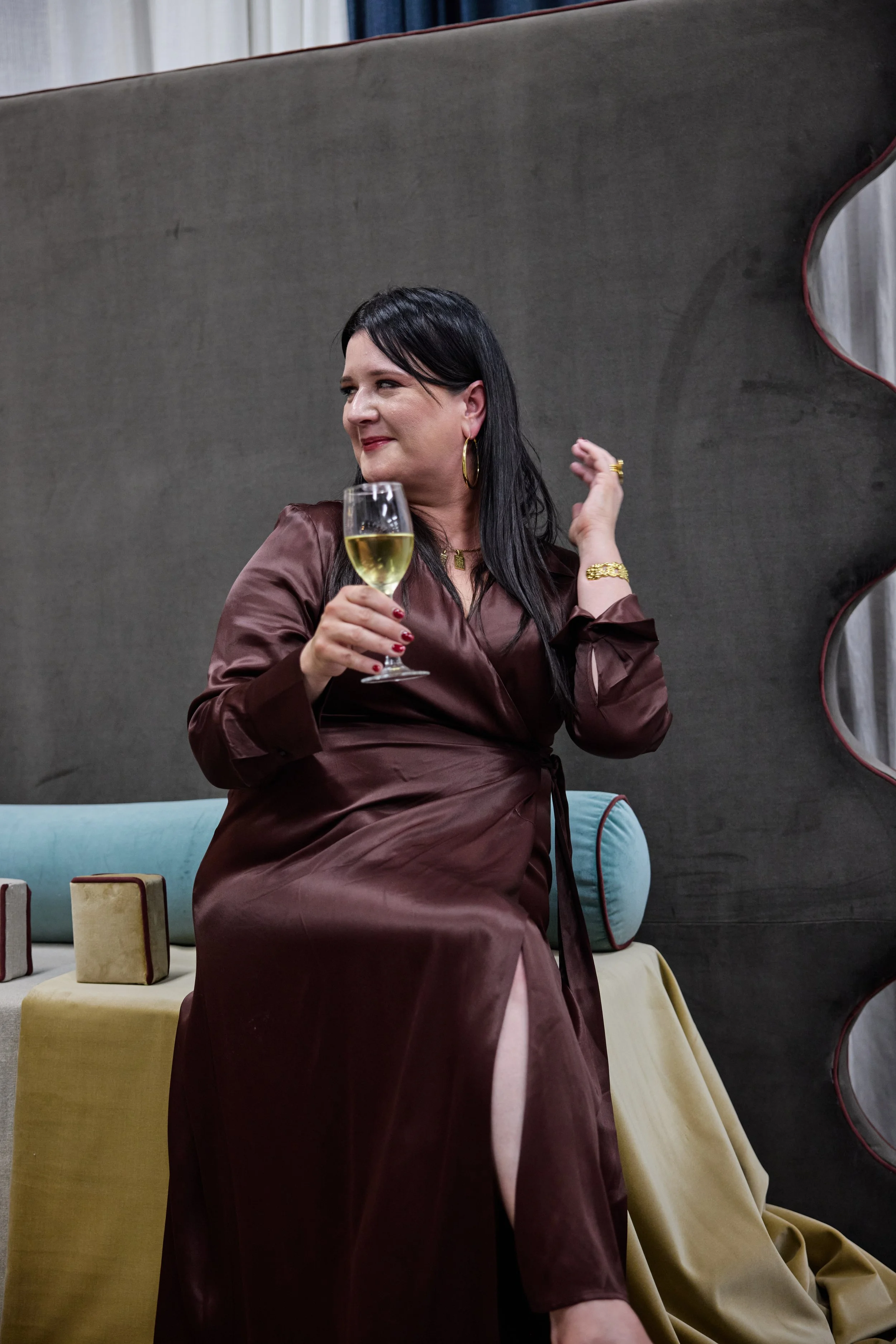 Woman in satin dress holding a glass of white wine, sitting on a sofa with decorative pillows.