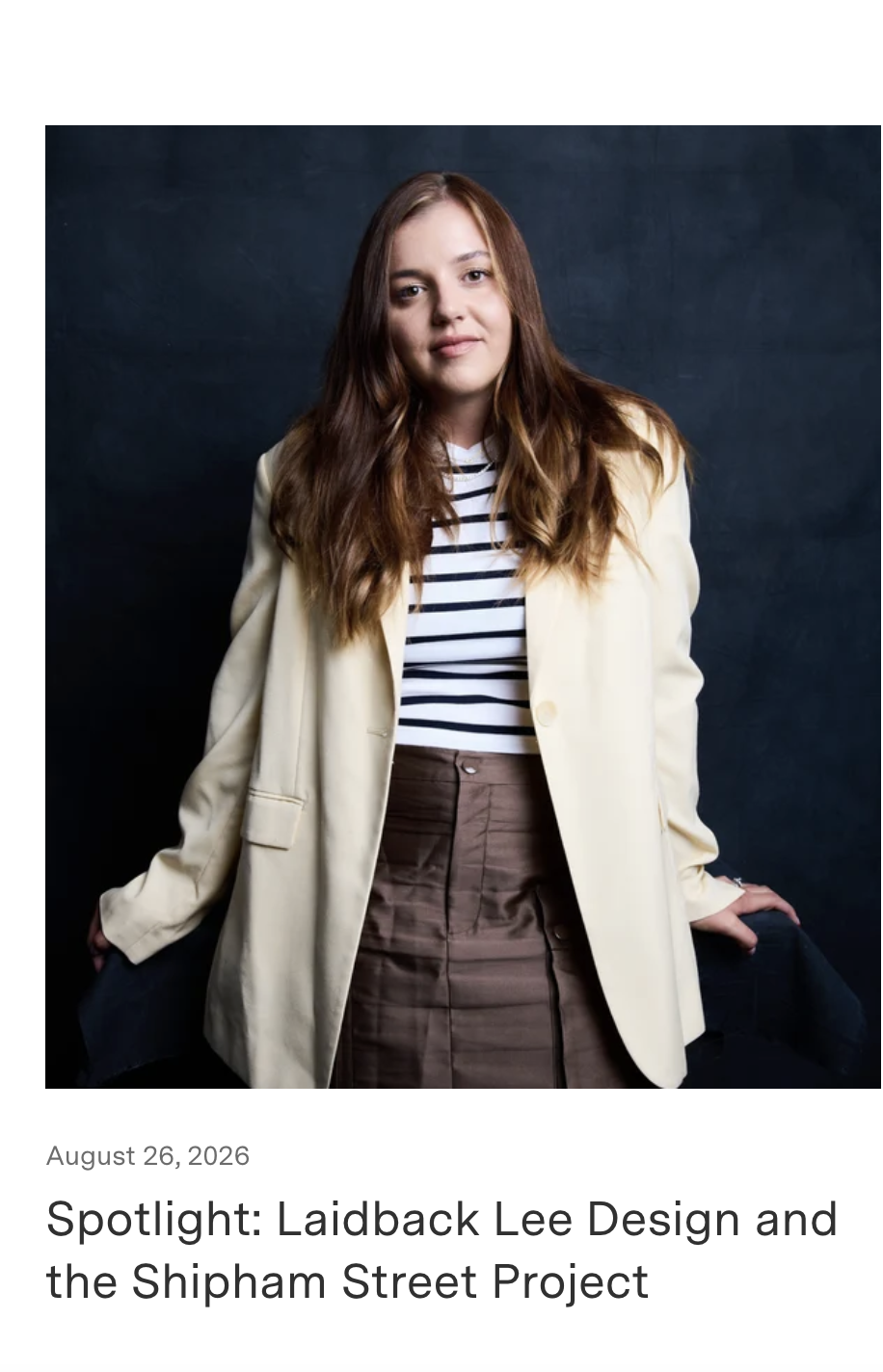 A woman with long, wavy brown hair wearing a long cream blazer over a black and white striped shirt and high-waisted brown pants, standing against a dark background.