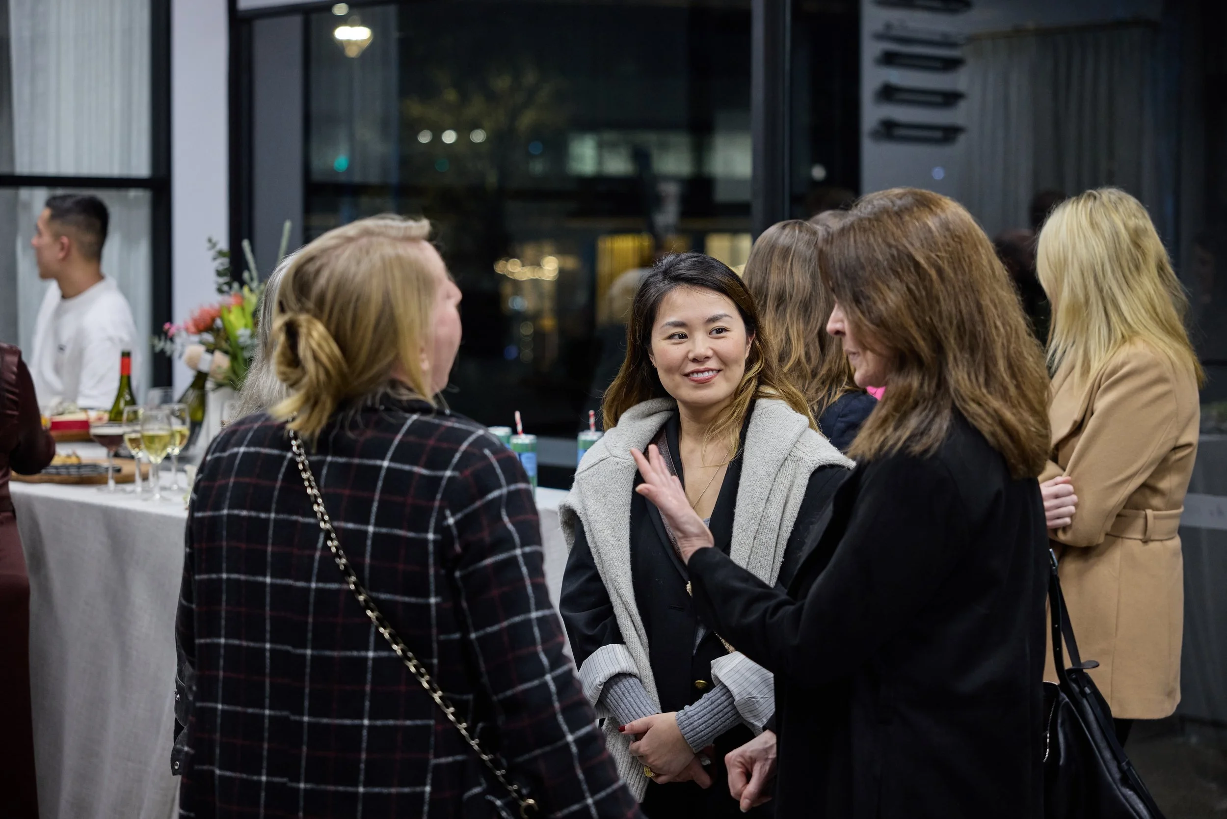 Group of women engaged in conversation at a social gathering indoors, with a table of drinks and food in the background.