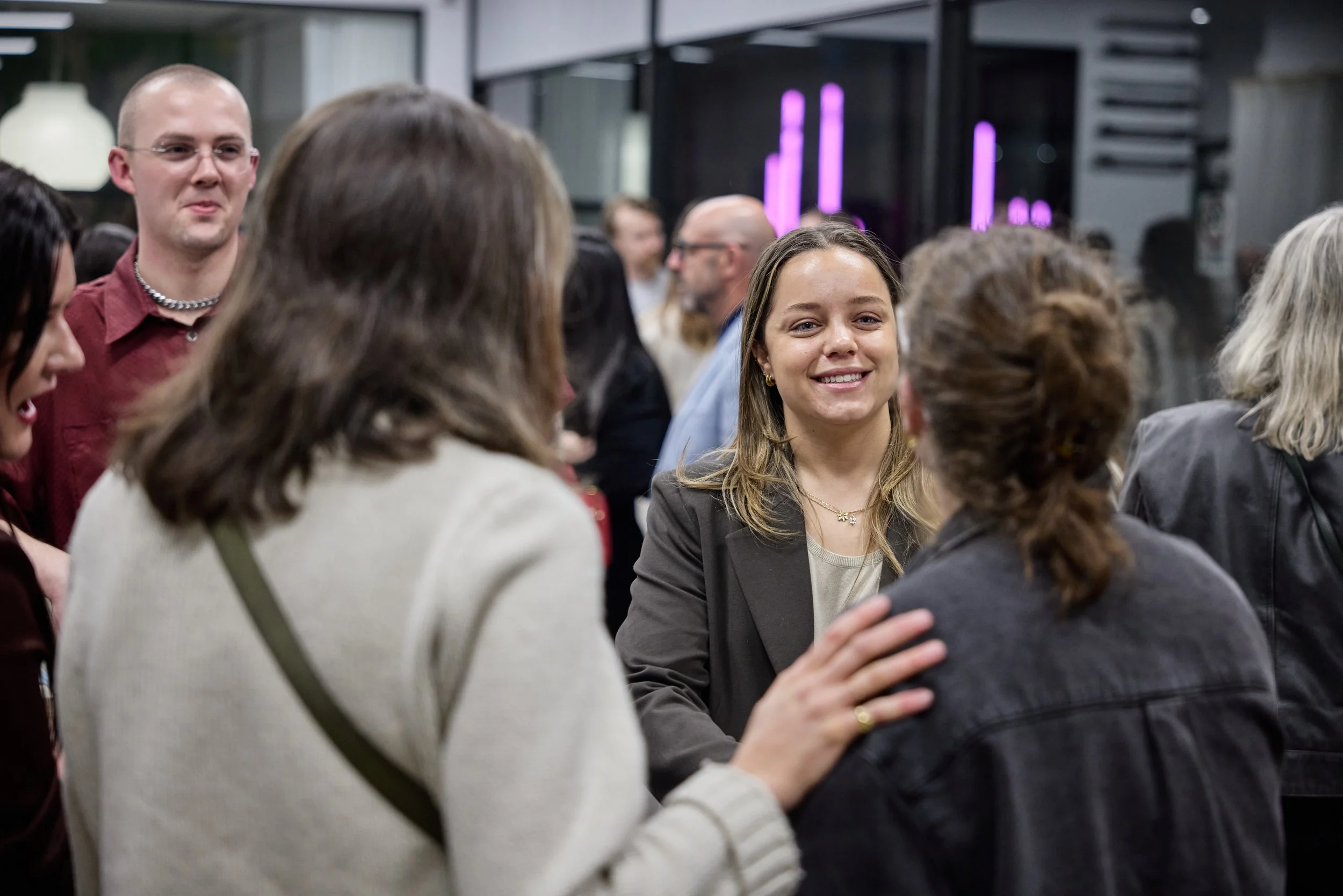 Group of people at a social event, conversing and smiling, with a young woman in a blazer and gold necklace in the center.
