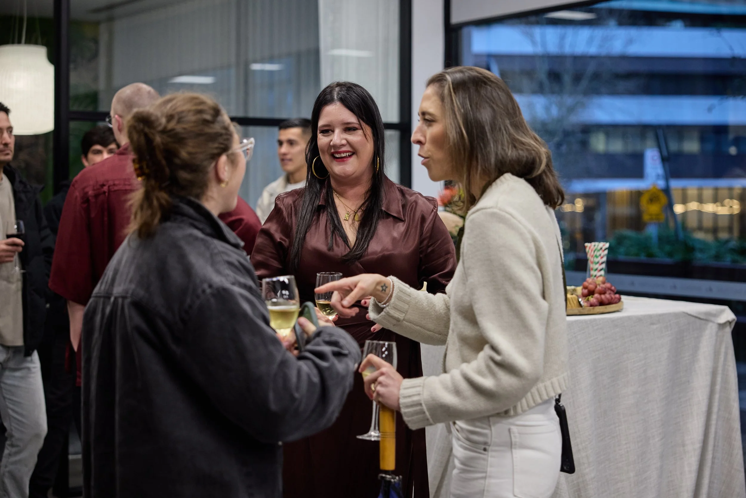 Group of women at a social gathering, chatting with drinks in hand, with a table of snacks and large window in background.