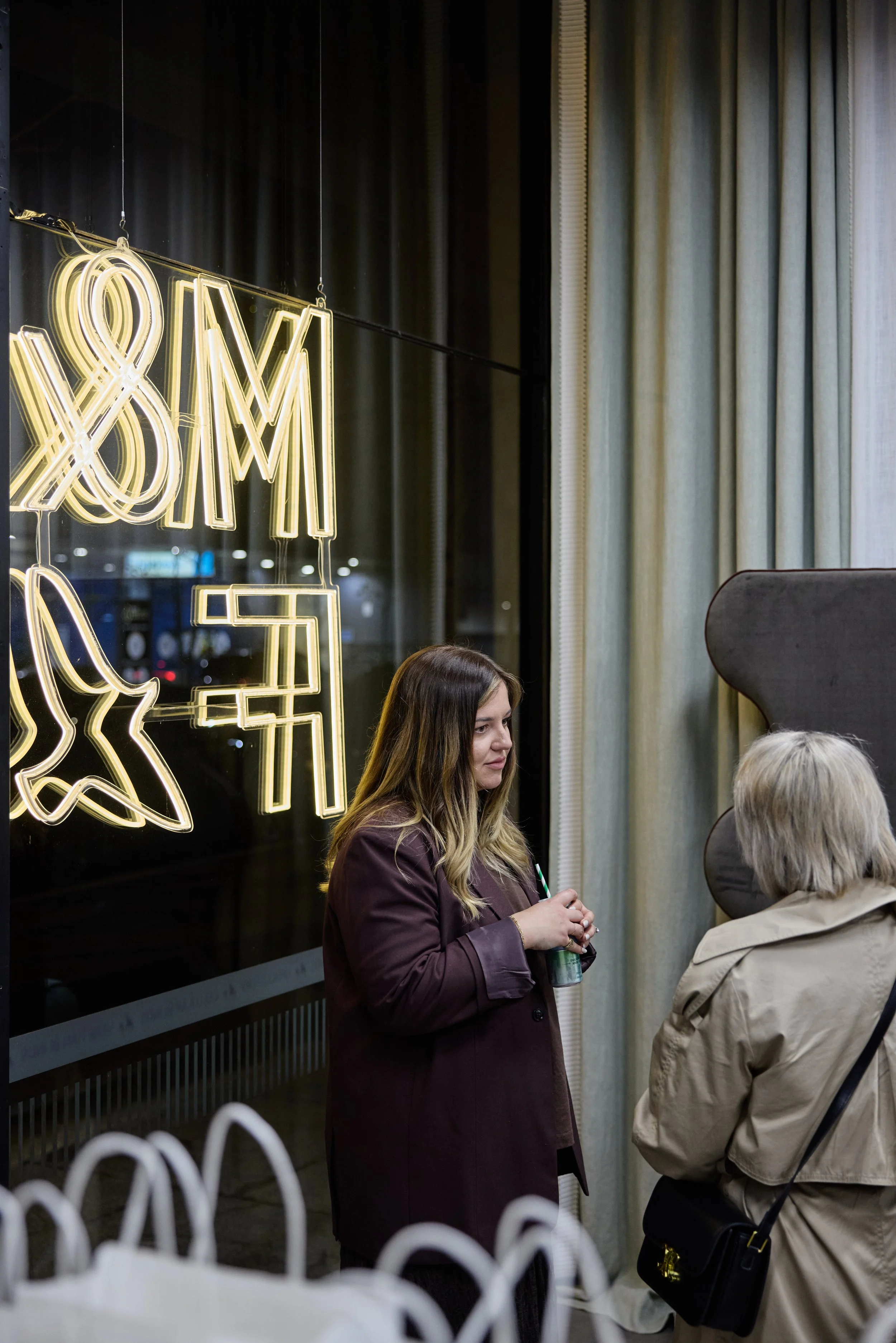 Two women are standing and talking in front of a large window with neon yellow decorative signs behind them, inside a modern building at night. One woman has long brown hair and is wearing a maroon coat, holding a drink. The other woman has short gra