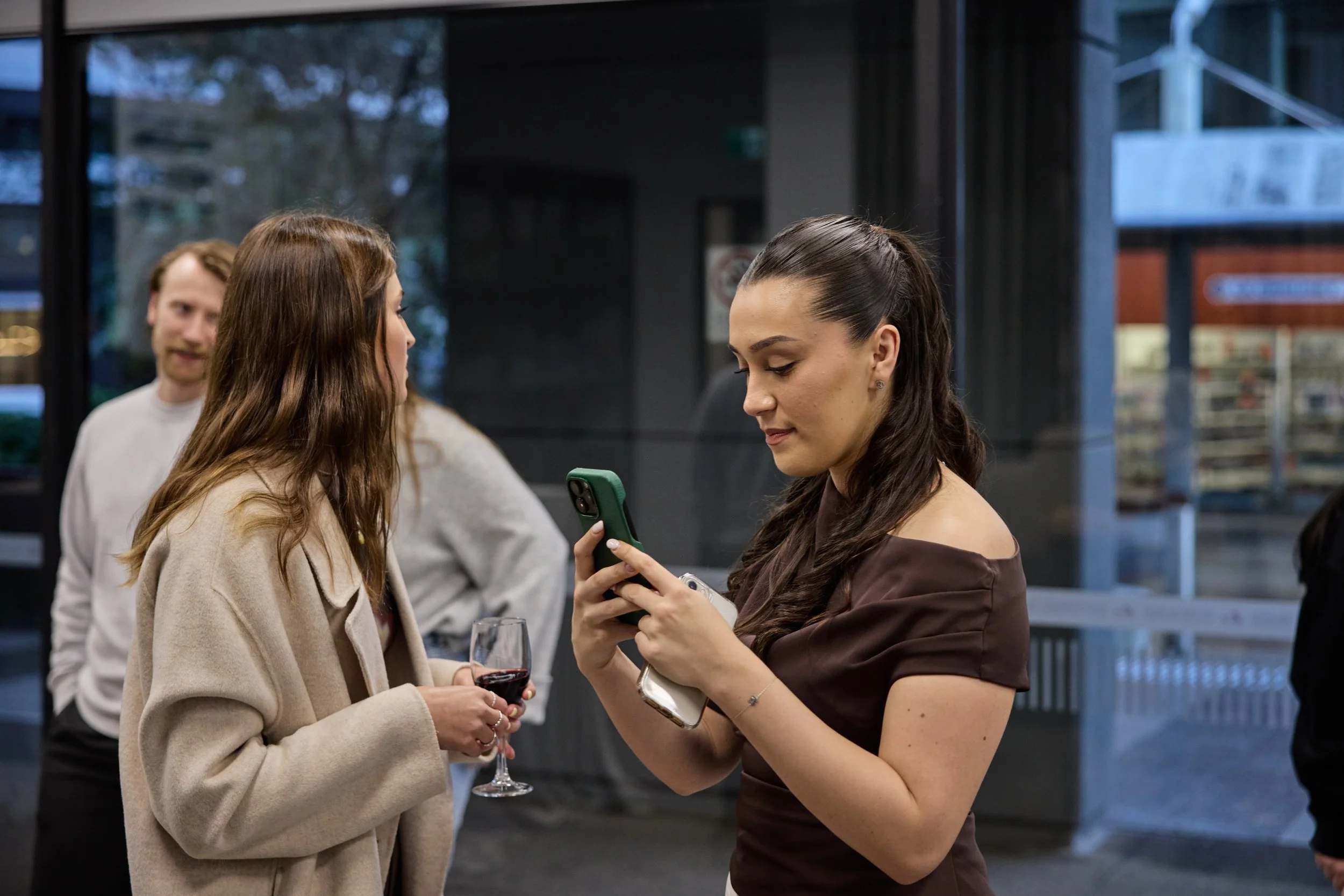 Three women at an indoor event, one of whom is holding a glass of red wine, and another is looking at her phone; a man is in the background.