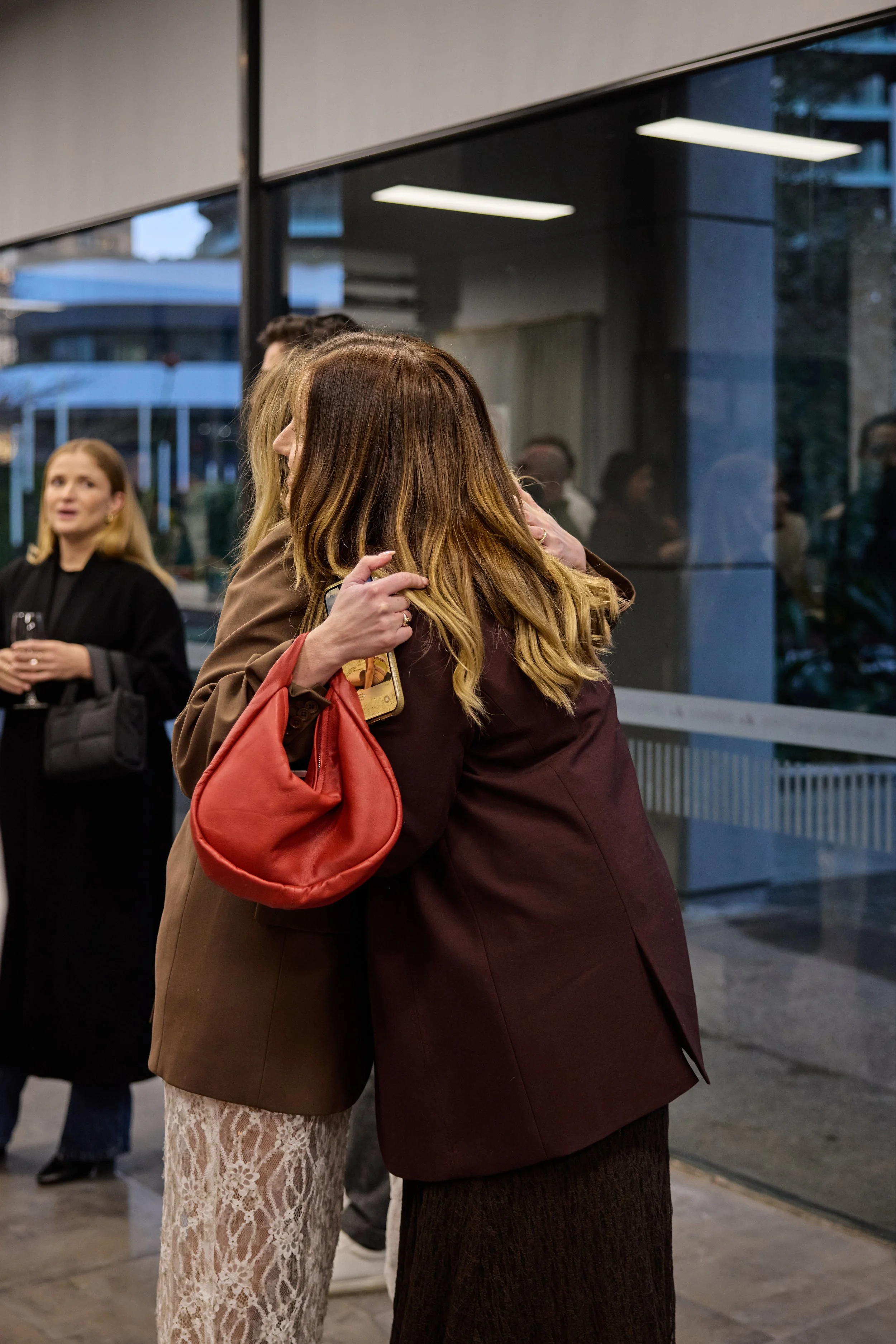 Two women hugging in a social gathering inside a building with large glass windows, with other people in the background.