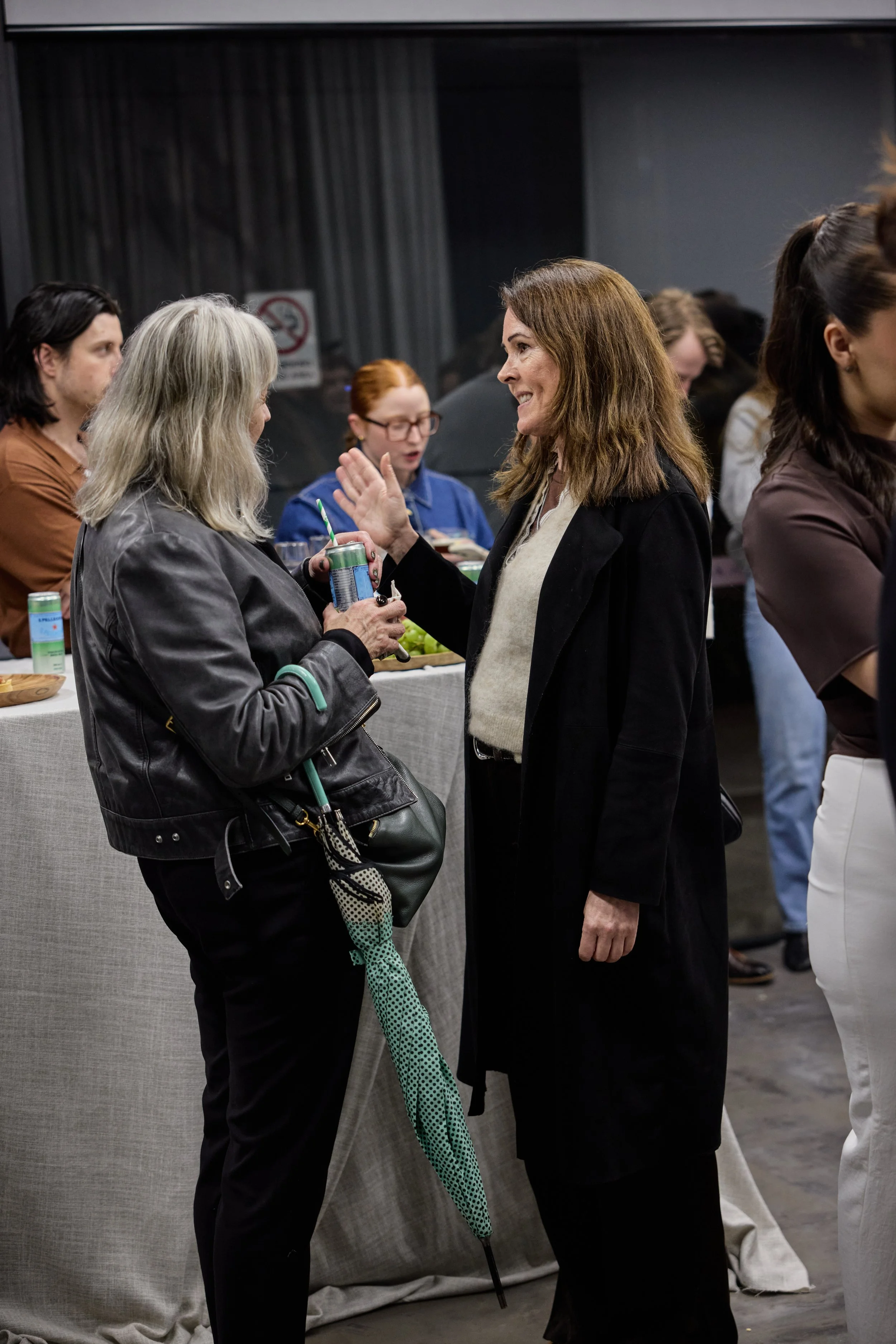 Two women having a conversation at a social event with others in the background behind a table with food and drinks.